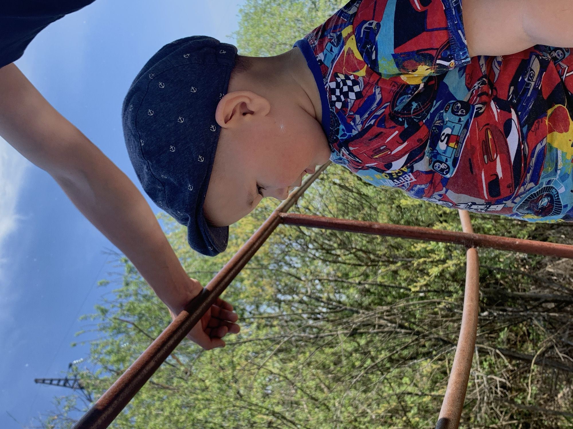 A child in a car-themed shirt and a blue cap with anchor patterns is positioned on a rusted metal structure outdoors. An arm, likely belonging to an adult, is also visible. The location is Фоки, Russia, identifiable by the presence of a partially visible industrial crane, an element common in urban landscapes in Russia. The setting includes greenery, with trees and foliage in the background.