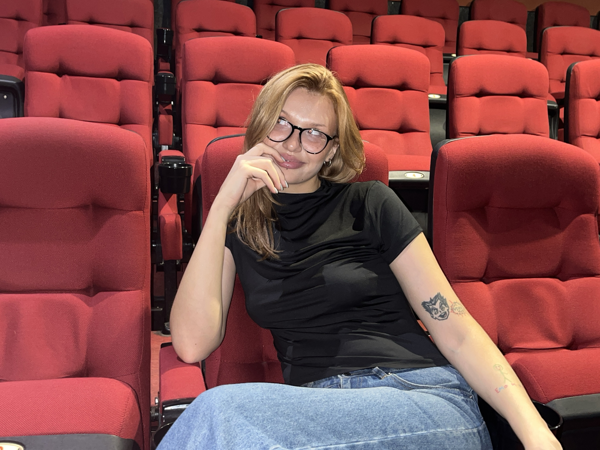 A young woman with blonde hair and glasses sits in a movie theater, resting her chin on her hand. She wears a black shirt and blue jeans. Her left arm, visible on the right side of the image, has two tattoos: a cartoon character on her upper arm and a stick figure with the word "Emo" written below it on her forearm. The seats are red, and some have cup holders.