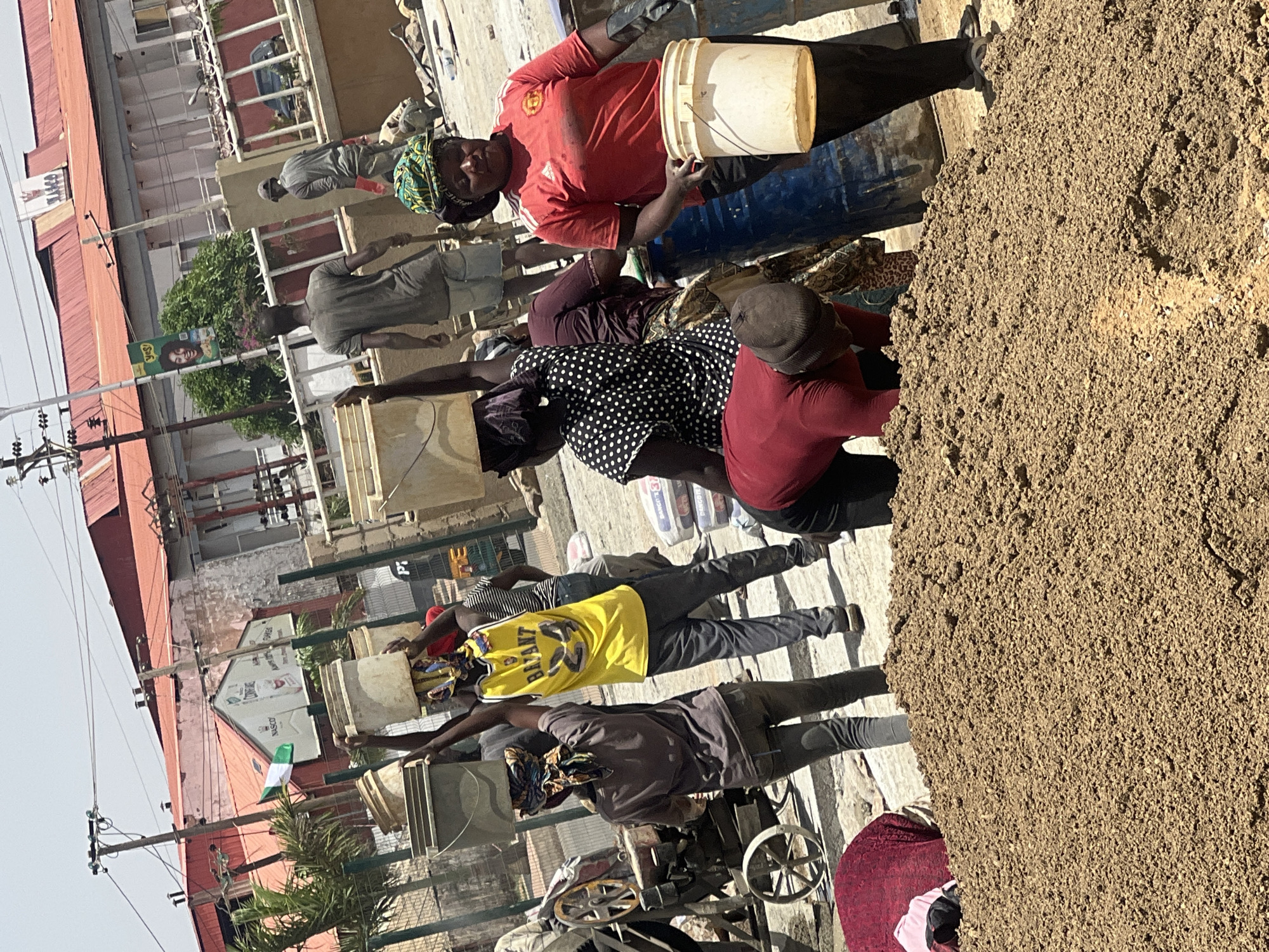 A group of people are carrying buckets, possibly of concrete, at what appears to be a construction site. The location is suggested to be Barakin Lerawi, Nigeria. The individuals are wearing various clothing items, including t-shirts and head coverings. In the background, there are buildings, street signs, and power lines, suggesting an urban environment. A pile of sand or gravel is visible in the foreground.