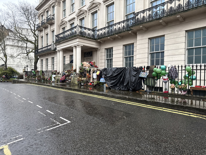 The image displays a wet street scene in front of a large, multi-story neoclassical building in London, United Kingdom, likely situated on Kensington Palace Gardens. The structure features a light-colored stucco or stone facade, numerous windows with dark frames, ornate black iron railings on balconies, and a prominent entrance portico with columns. The sky is overcast.
The asphalt road surface is wet, showing reflections, indicating recent rain. Double yellow lines run along the curb, and white dashed lane markings are visible on the road. Along the entire length of the building's black iron fence, a substantial public memorial is arranged. This memorial includes numerous bouquets of flowers, various soft toys and teddy bears, green balloons tied to the railings, personal notes, small flags, and other commemorative objects. A significant section of the fence is covered by a large black tarpaulin. Garments, including a striped top, are draped over parts of the fence. A small wooden bench or platform on the far right also holds additional memorial items. Bare trees are visible to the left of the building, and a dark lamppost stands near the sidewalk. No individuals are present in the scene.