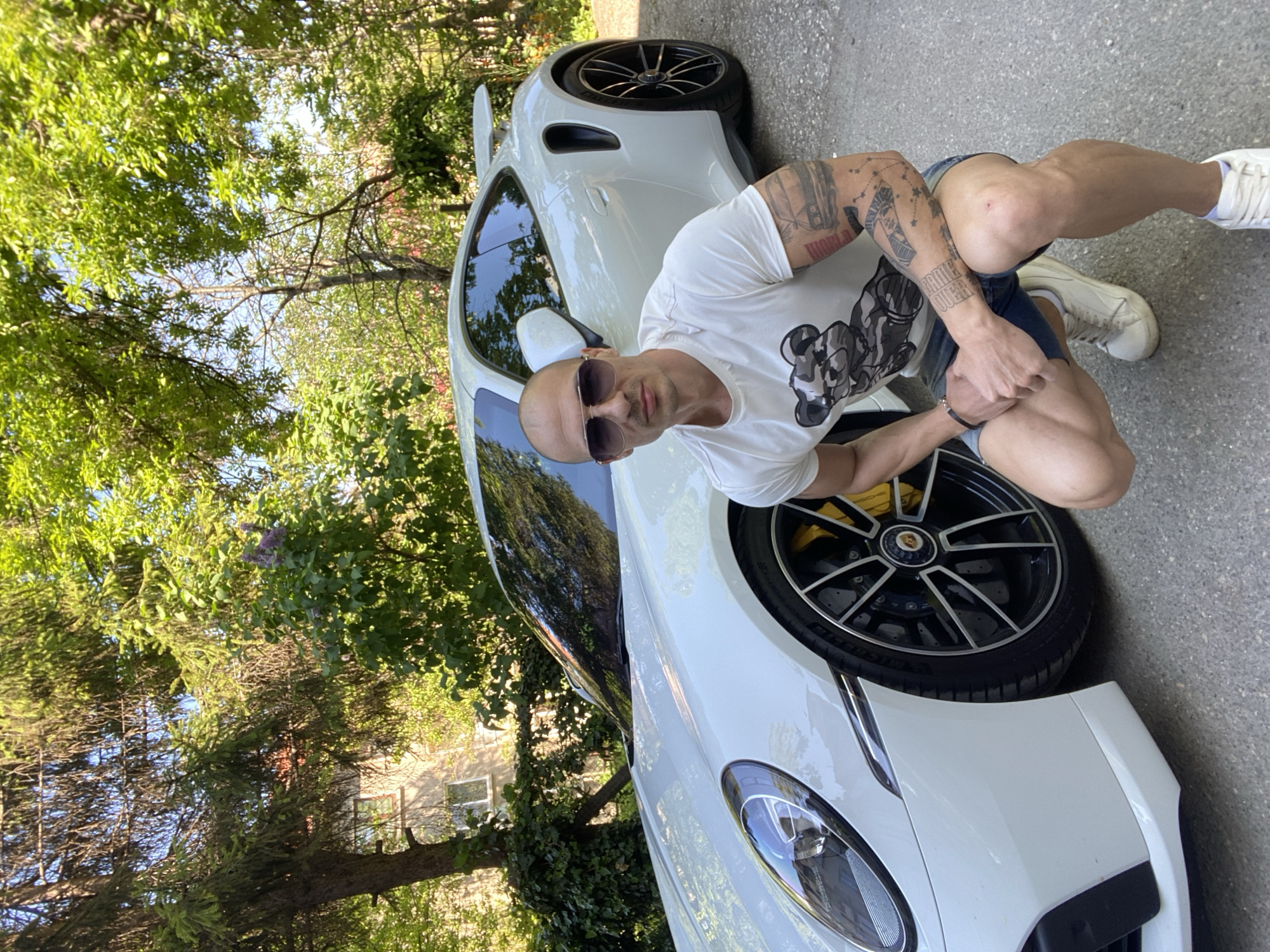 A man with tattoos is crouching next to a white Porsche. The background includes trees and a building. The location is Pleven, Bulgaria.