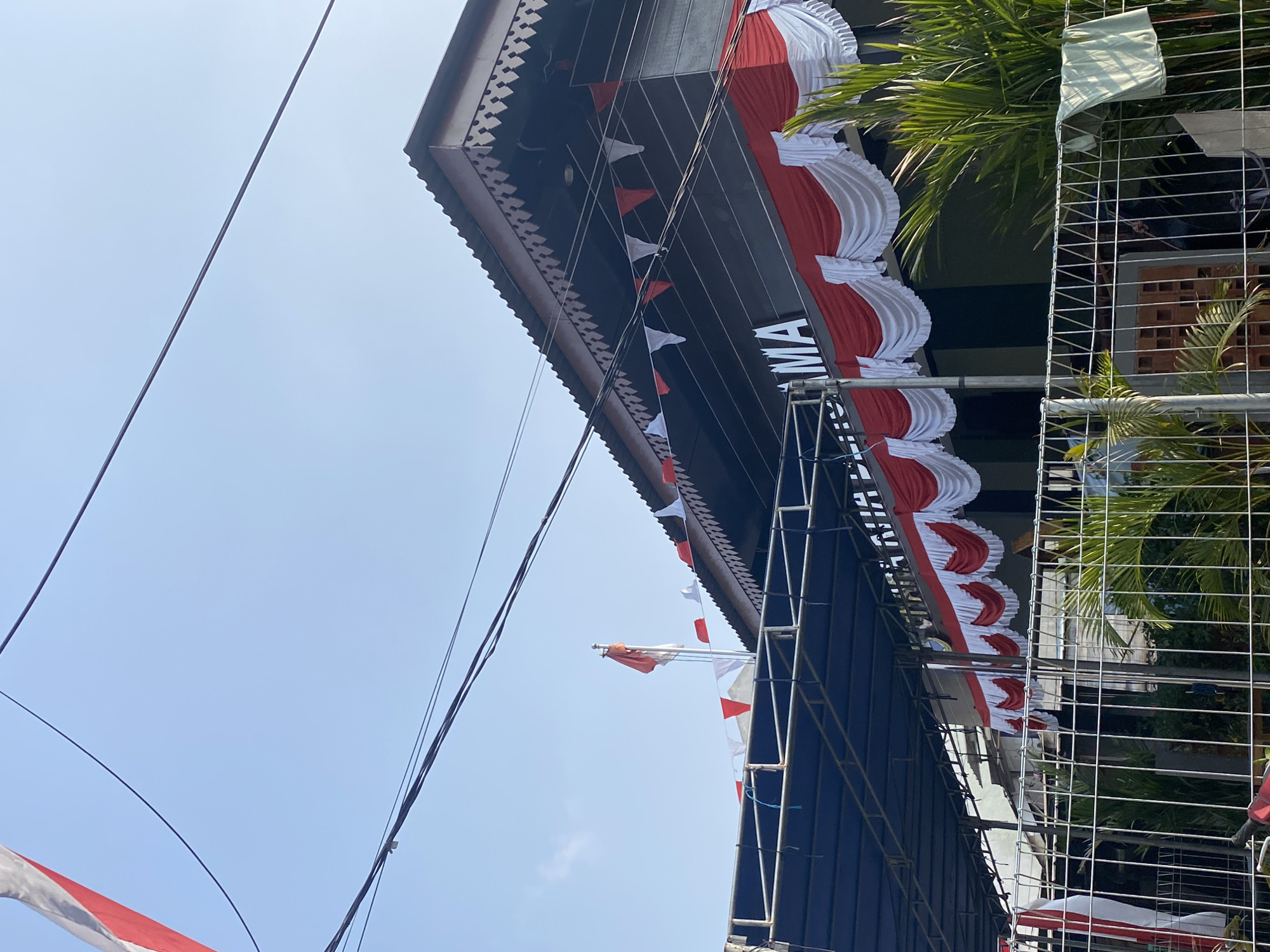 The image shows a building with red and white festive decorations, likely for a national celebration, possibly in Tangerang, Indonesia. A flag, appearing to be the Indonesian flag with its red and white colors, is visible on a flagpole atop the building. The building has a dark facade and a corrugated metal roof with decorative trim. Red and white bunting or fabric is draped along the edge of the roof and also hangs in swags above a covered walkway. Small red and white triangular flags are strung along a wire. Overhead power lines crisscross the sky. In the foreground, a metal fence encloses an area with some greenery, including palm fronds. A light-colored piece of fabric is hanging on the fence. The sky is clear and blue. The overall impression is one of a public or community building decorated for a festive occasion.