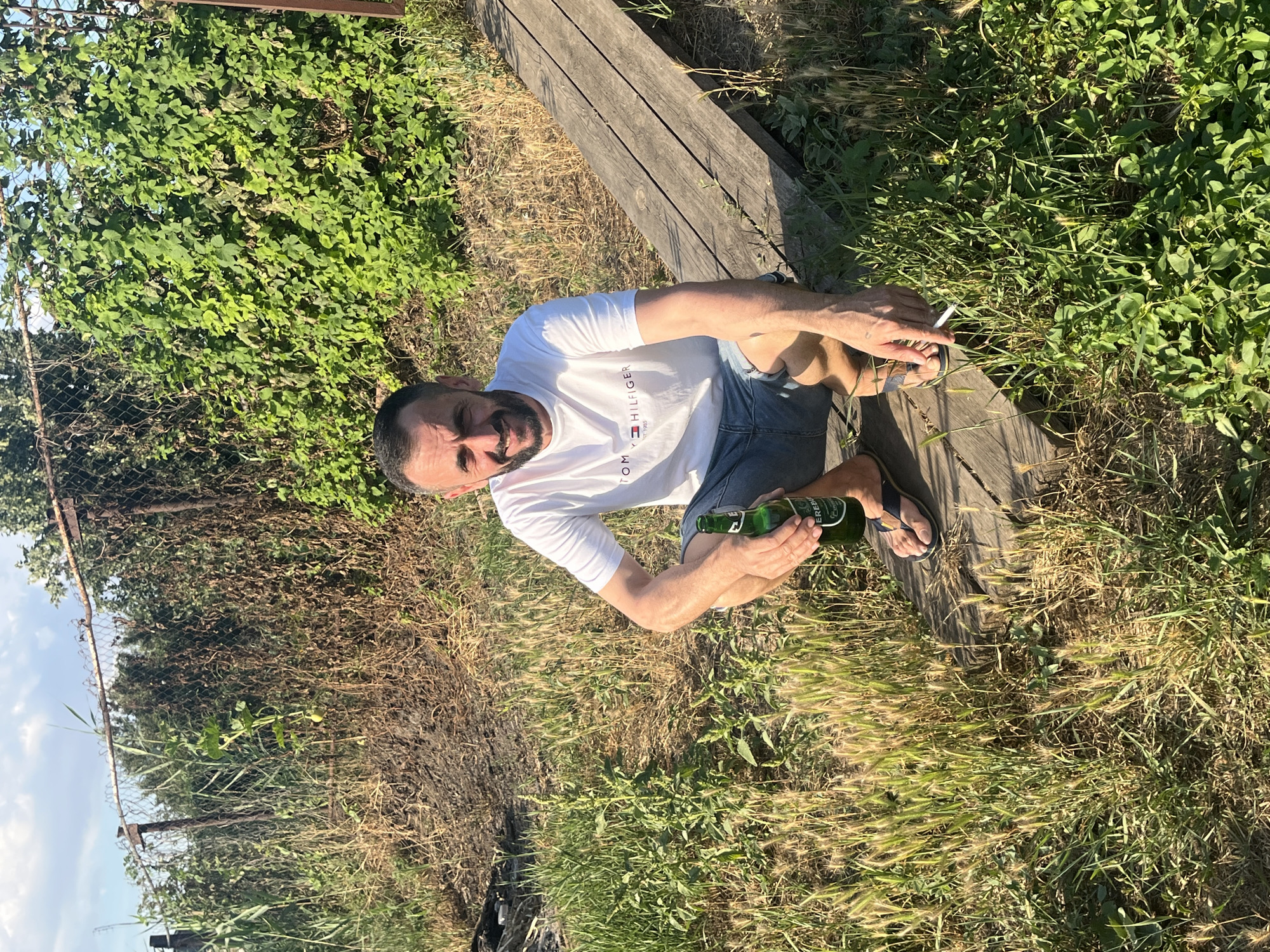 A man with dark hair and a beard is crouching on a wooden plank structure outdoors. He is wearing a white Tommy Hilfiger t-shirt and denim shorts. In his right hand, he holds a green bottle of beer, and in his left hand, he holds a lit cigarette. The background consists of a wire fence, overgrown vegetation, and a clear sky with some clouds. The ground is covered in grass and dry vegetation. The scene appears to be in a rural or semi-rural setting, possibly a garden or backyard. The specific location in the city of Яново-Грушевский, Russia, cannot be determined from the image alone.