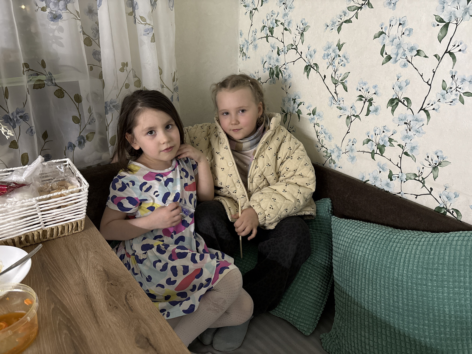 Two young girls are sitting on a brown sofa in an indoor setting. The girl on the left has dark brown hair, fair skin, and is wearing a colorful dress. She is looking towards the camera. The girl on the right has blonde hair styled in pigtails, fair skin, and is wearing a beige quilted jacket over a patterned top. She is holding a thin stick-like object.
The room is decorated with floral wallpaper. To the left of the girls, there is a wooden table with a wicker basket filled with food items, a plate, a spoon, and a small container. Curtains with a floral pattern hang nearby. Green cushions are positioned behind the girls on the sofa.
There is no explicit information in the image to confirm that the scene is located in Chaikovsky, Russia.