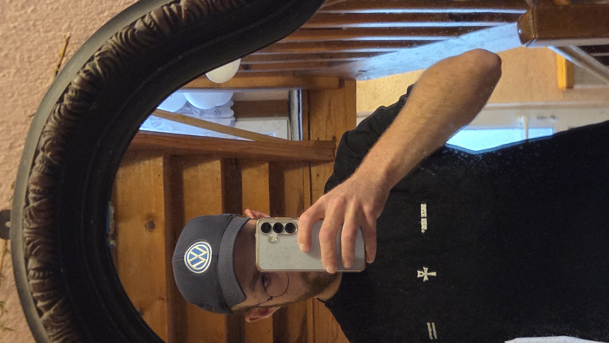 A person is reflected in a mirror, holding a smartphone to take a selfie. They are wearing a dark blue baseball cap with a white Volkswagen logo, round wire-frame glasses, and a black t-shirt with white graphic designs. The background of the reflection shows a wooden staircase and a door. The scene appears to be indoors, in a domestic setting.