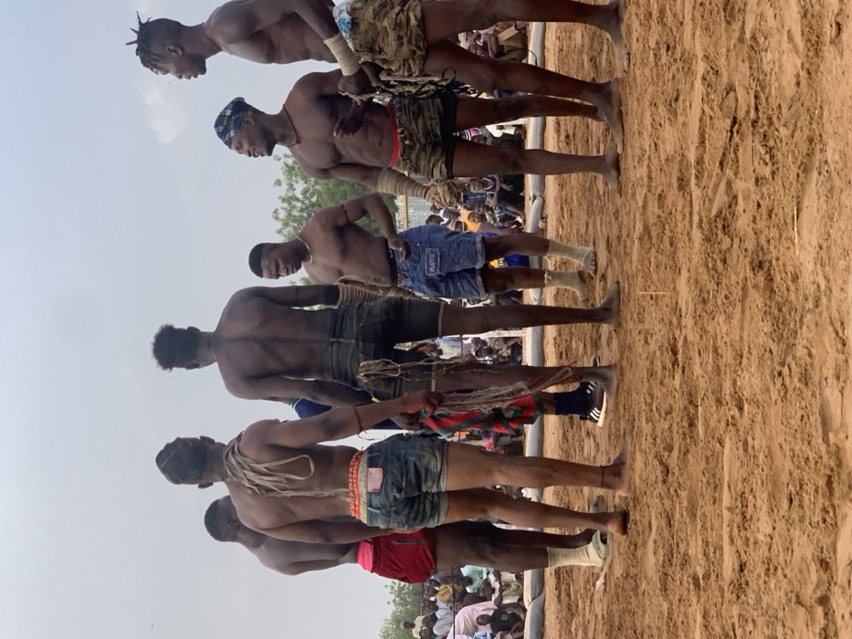 The image shows a group of six men standing on a sandy wrestling pit in Sokoto, Nigeria. They are all shirtless, wearing shorts and some have traditional head coverings. They are facing each other, but are not actively engaged in wrestling. The scene is likely a pre-match preparation or a break during a wrestling tournament. Several people are seated in the background as spectators, watching the men. The sky is overcast, but it is daylight. The coordinates provided place the scene in Sokoto, a city in northwestern Nigeria. The environment is a sandy arena, likely an outdoor wrestling ground.