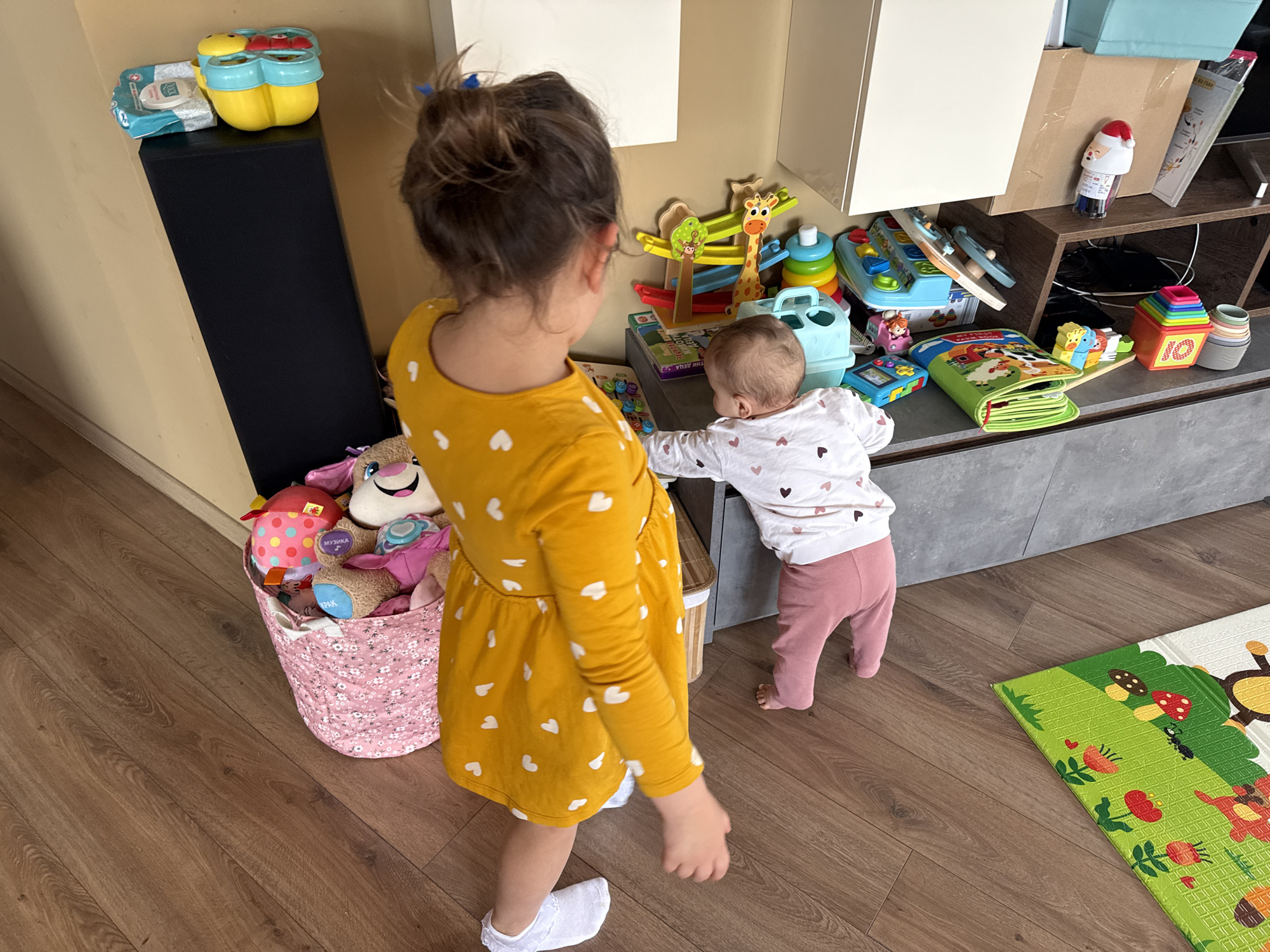 The image shows two young children in an indoor setting. The elder child, a young girl with brown hair in a bun, wears a yellow dress adorned with white hearts and white socks. She faces away from the camera. A younger child, appearing to be a baby, wears a white long-sleeved shirt with hearts and pink leggings. This baby is reaching toward a surface covered with various toys. These toys include a giraffe-themed toy, a blue toy toolbox, a green book, and colorful stacking cups. Behind these items is a light gray TV stand, partially obscured by a box with a Santa Claus figurine on top. The room features a black speaker, a pink fabric basket overflowing with plush toys, and a foam play mat with a jungle scene on the wooden floor. The location is inferred to be indoors, potentially in a home or daycare center in Sofia, Bulgaria.