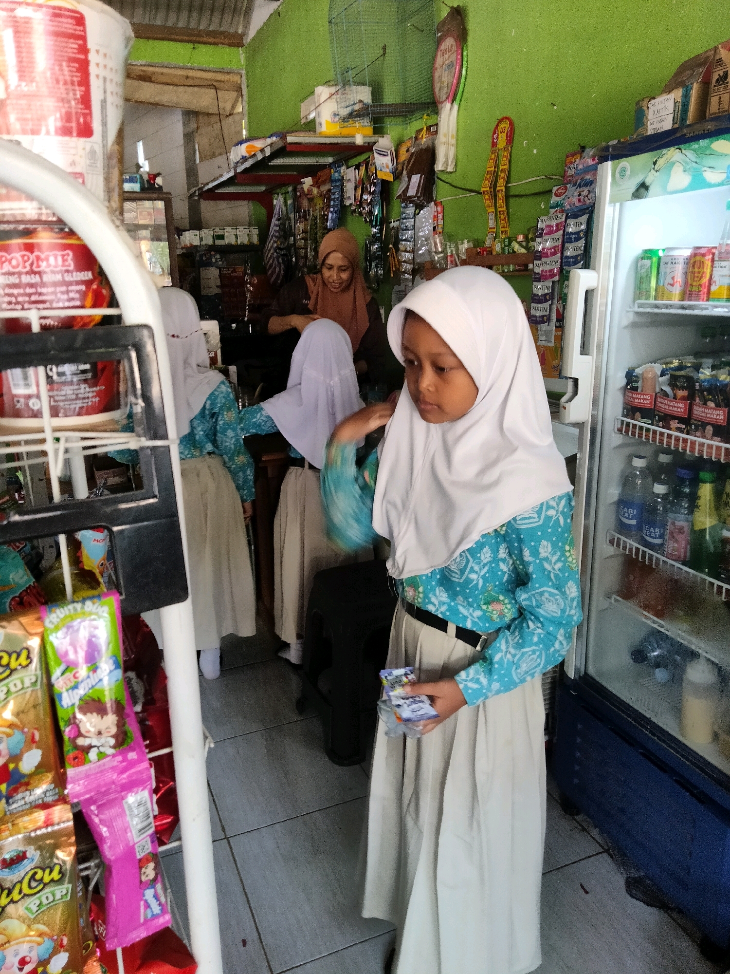 In a dimly lit convenience store with green walls, a young girl wearing a white hijab and a blue and white patterned tunic stands in front of a blue refrigerator. She holds a small packet of candy in her hand. Behind her, two other girls in similar attire are partially visible, with one in the background looking at the camera. Shelves stocked with various products, including drinks and snacks, line the walls. A metal display rack with packaged snacks is to the left of the frame. The floor is tiled with gray squares.