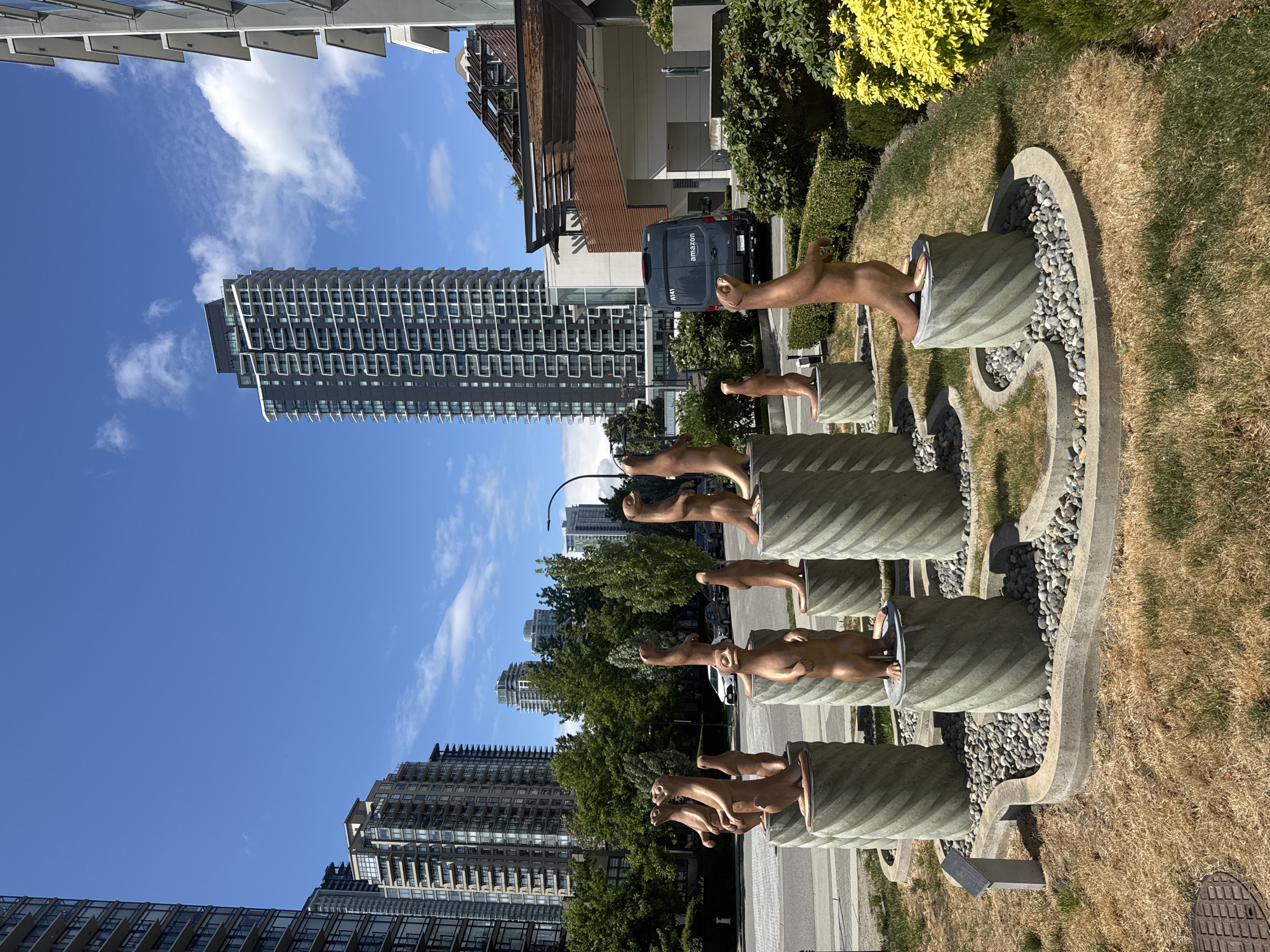 The image shows a public art installation featuring bronze sculptures of otters standing on cylindrical pedestals. The sculptures are arranged in a semi-circular formation on a bed of river stones, bordered by a concrete curb. The foreground features dry, brown grass and a section of a manhole cover. In the background, a modern city skyline is visible, with tall apartment buildings and a darker, modern skyscraper dominating the view. A dark-colored Amazon delivery van is parked near a building with a modern architectural design featuring wooden elements and a covered outdoor area. The sky is a clear, bright blue with scattered white clouds. The scene appears to be located in an urban park or plaza area in Burnaby, Canada, given the architectural style of the buildings and the presence of public art.