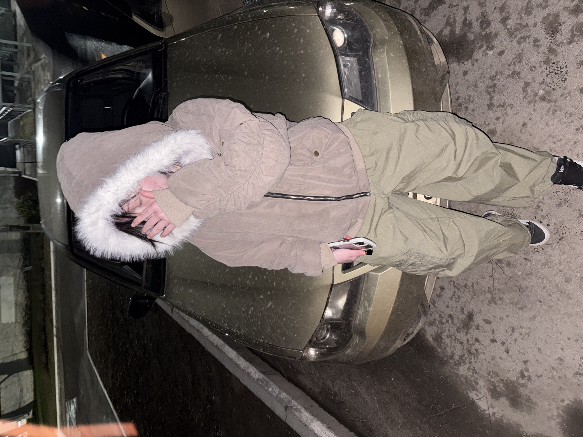 The image, taken in Chaikovsky, Russia, captures a person lying prone on the dirty, light-colored hood of a car at what appears to be nighttime or late evening. The individual is dressed in a thick, light brown or beige winter jacket with a prominent white faux fur-lined hood, and olive green baggy pants. Their face is largely obscured by the hood and their hands, which cover the lower part of their face, though dark hair is visible. A small star tattoo is visible on the back of one hand. They are holding a white smartphone with a dark case in their right hand. Their feet are clad in black and white Vans-brand sneakers.
The car, an SUV or crossover vehicle (possibly a Nissan Pathfinder, given the grille and headlight design), is noticeably dusty or speckled with dirt and possibly salt/slush, suggesting recent exposure to damp or cold conditions. The driver's side door is open. The ground beneath and around the vehicle is paved (concrete or asphalt) and appears wet or damp in various spots. The dim, artificial lighting and heavy winter clothing further indicate a cold, dark environment. The background is dark and indistinct, showing vague outdoor structures. The person's posture could suggest rest, weariness, or a casual pose for a photograph.