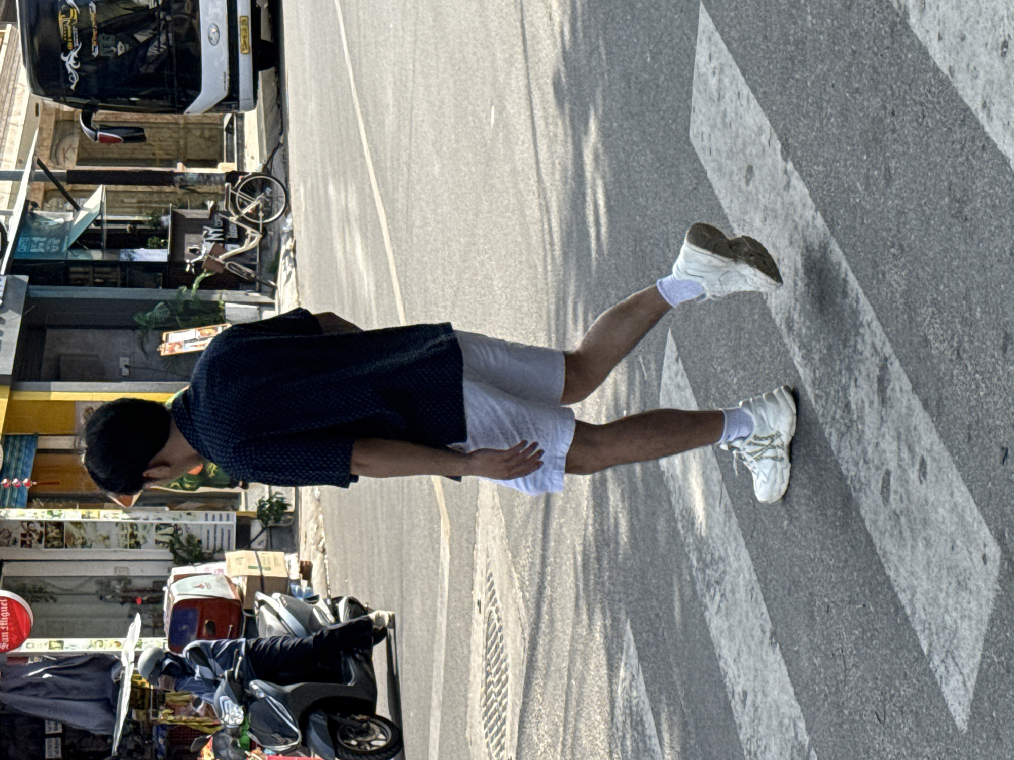 A person is walking across a street on a crosswalk. The person is wearing a dark patterned short-sleeved shirt and white shorts. They have on white sneakers with white socks. The background shows shops and a bus. The street appears to be in a city environment. The image was taken in Bac Nha Trang Ward, Vietnam.
