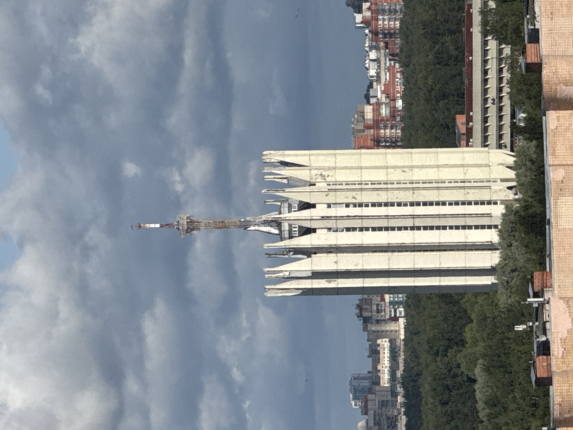 An outdoor, medium-distance, slightly high-angle shot captures a tall, futuristic-looking concrete building topped with a communications tower against a cloudy sky. The building features a unique design with pointed vertical fins running up its sides, framing a central section with narrow, rectangular windows. Below the main building, a dense, dark green forest stretches across the middle ground, interspersed with glimpses of other urban structures. In the background, a sprawling cityscape of residential and industrial buildings is visible under the overcast sky. The foreground shows the roof of a brick building with chimneys. The image appears to be taken in an urban environment, possibly St. Petersburg, Russia, given the architectural style and surrounding vegetation. There are no people visible in the image.