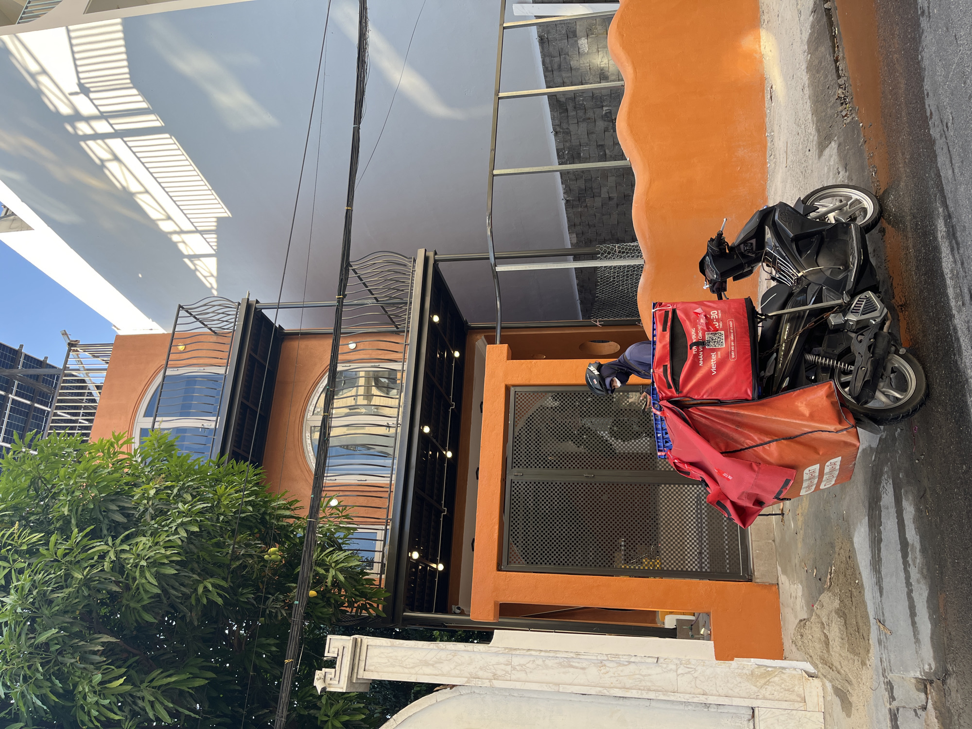 Here's a description of the image:
The photo shows a black moped parked on a street in front of a building with an orange facade. The moped has two delivery bags attached: a red Viettel Post bag and an orange bag with Vietnamese text. A person wearing a blue jacket and helmet is sitting on the moped. The building features large arched windows with reflective glass, and the structure is partially obscured by a leafy tree. Wires stretch across the top of the frame. The building appears to have a contemporary design, with metalwork on the exterior. A marble-like wall runs along the bottom of the frame. The location is suggested to be Đà Nẵng, Vietnam. The street appears to be partially wet.