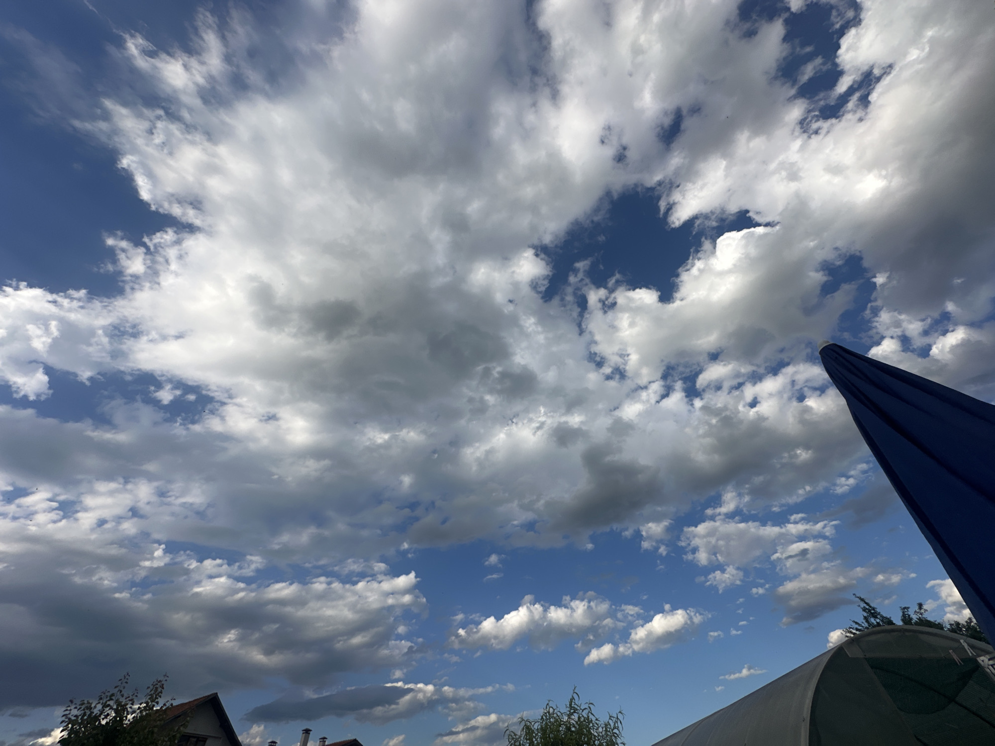 The image shows a partly cloudy sky with the sun peeking through. The clouds are a mix of white and gray, with some darker, heavier clouds suggesting possible rain in the distance. The sky is a vibrant blue, indicating a clear day despite the clouds. In the foreground, there's a glimpse of rooftops of houses, characteristic of the village of Cherven Breg, Bulgaria. A large dark blue umbrella is visible on the right side, suggesting an outdoor setting, possibly a garden or patio area. The overall scene depicts a typical rural landscape under a dynamic sky.