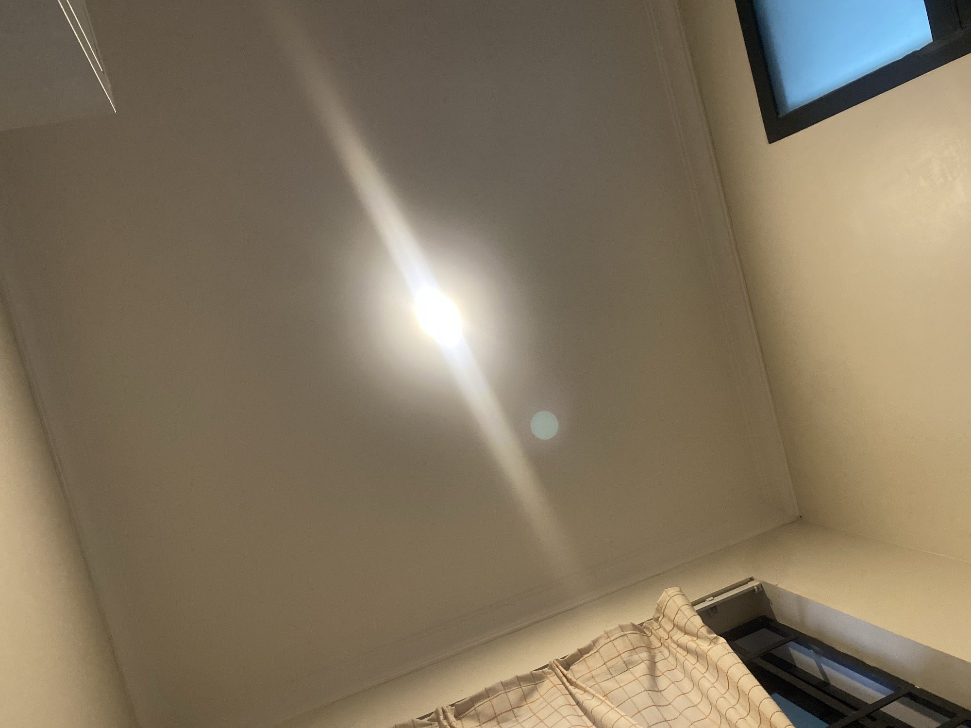 The image displays an upward view within an interior room, focusing on the ceiling and upper sections of the walls. A prominent, bright artificial light source, possibly an LED fixture, is centered on the off-white ceiling, emitting an intense, elongated beam of light and a circular lens flare. The walls are painted a light beige or cream color. In the top right corner, a dark-framed window or opening reveals a clear, bright blue sky, suggesting it is daytime outdoors. Another dark-framed window or opening is visible in the bottom right, partially obscured by a light-colored, checkered curtain. A white fixture, possibly an air vent or part of the window frame, is located above this lower window, behind the curtain. The scene is static, devoid of people or activity. The room appears simply furnished, illuminated by both the artificial ceiling light and natural daylight entering from the windows. No text is visible in the image. The location context is Dar es-Salaam, Tanzania.
