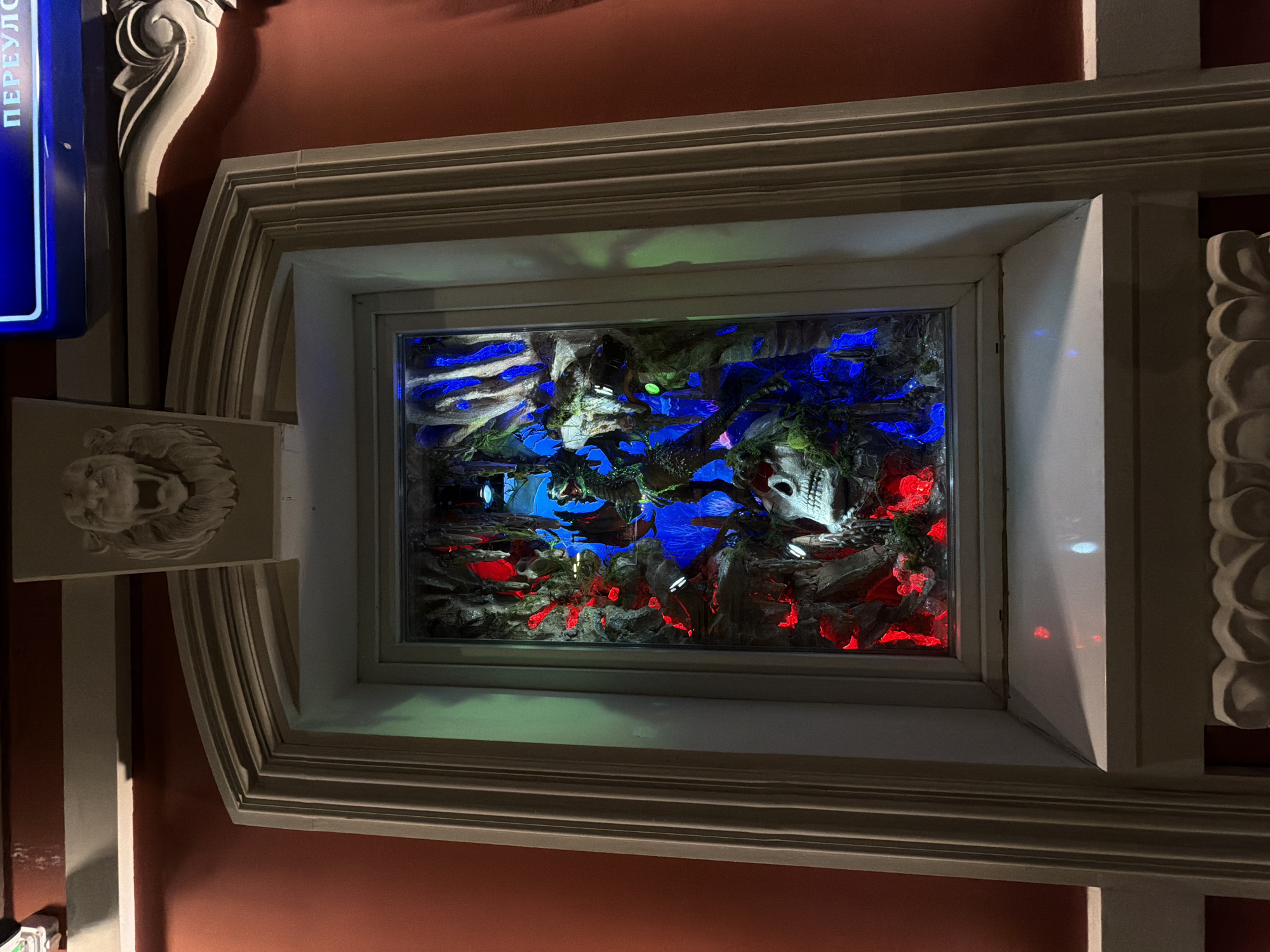 Here is a detailed description of the image:
The image shows an elaborate, framed display case containing a diorama or artistic arrangement. The frame is ornate, with layered moldings and decorative elements, including a lion's head relief on one side and a fluted column detail on the other. The diorama within the case features a scene lit with blue and red lights, depicting rocks, possibly skulls, and vegetation covered in moss. The overall setting appears to be indoors, with a reddish-brown wall visible in the background.
The location is Moscow, Russia.