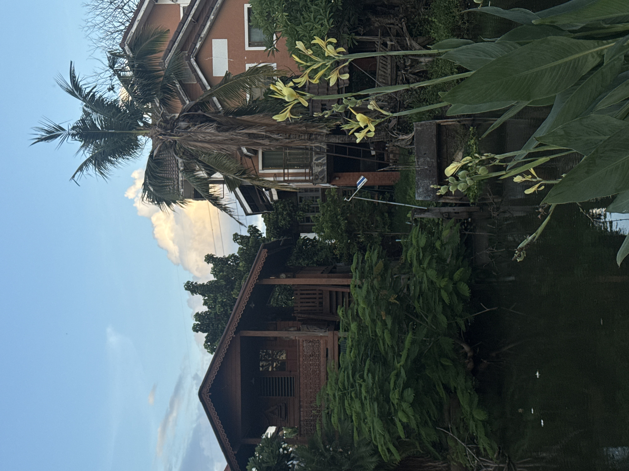 The image depicts a serene waterside scene at Baan Kanchanok 23 in Thailand. The setting features a calm body of water reflecting the surrounding lush greenery and a two-story building with an orange facade. In the foreground, a wooden house with intricate carvings on its facade is nestled amongst dense foliage. Yellow flowers bloom on tall stalks, adding a touch of color to the scene. The sky is a clear blue, with a few wispy clouds, suggesting it is daytime, likely late afternoon given the soft light. The overall mood is peaceful and tranquil, evoking a sense of tropical relaxation. There is no visible text in the image.