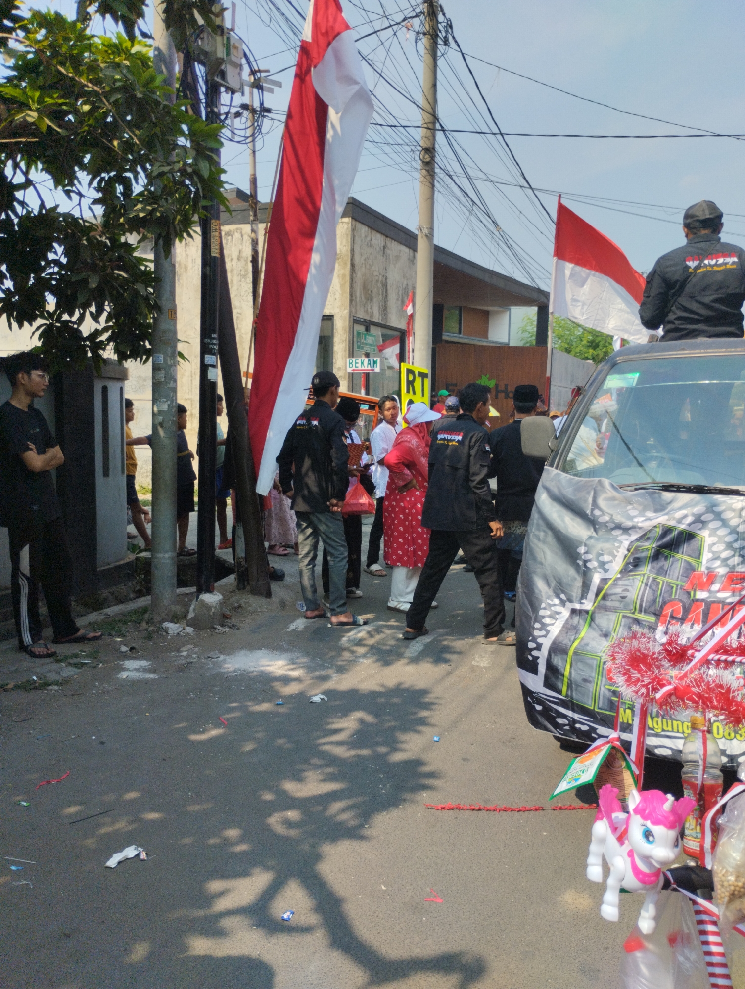 In Bekasi, Indonesia, on a sunny day, a group of individuals are gathered on a street. Several people, some wearing black shirts with logos and others in traditional Indonesian attire, stand around. Red and white Indonesian flags are prominently displayed, one large one hanging from a pole and another smaller one on a vehicle. The street is asphalt and appears somewhat littered with small pieces of red string and white debris. A black vehicle, decorated with a large banner featuring graphics and text, is parked on the right side of the image. On the vehicle's hood, a pink and white toy unicorn is visible, attached with red and white ribbons. The background shows buildings and utility poles with numerous wires. The overall atmosphere suggests a local event or celebration taking place outdoors.