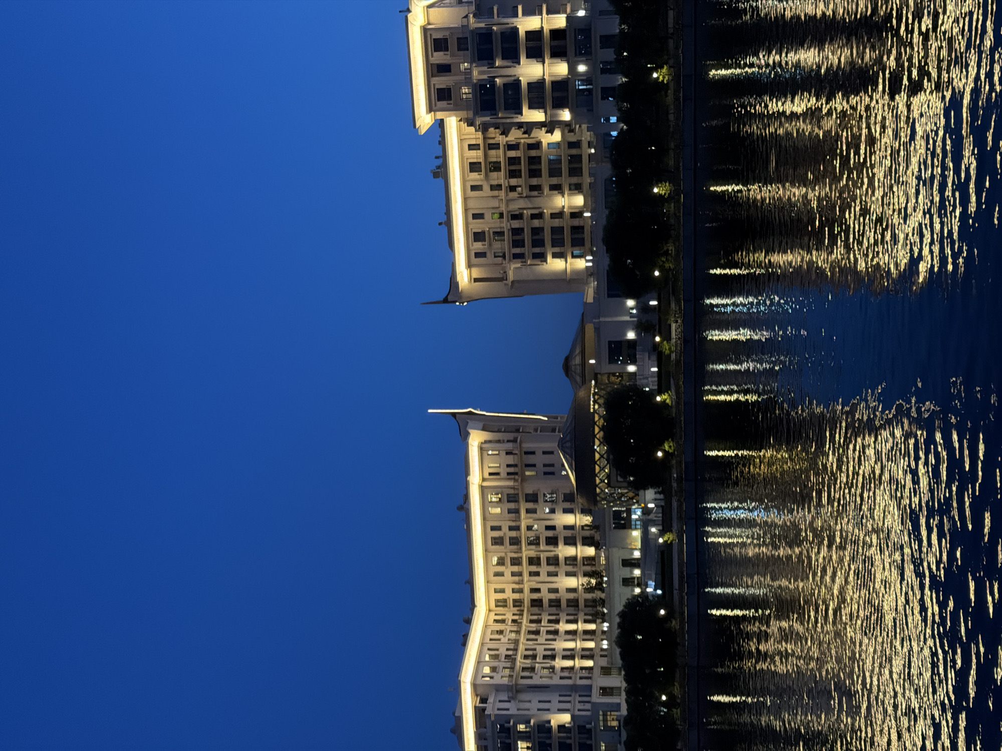 The image depicts a twilight scene of modern buildings reflected in a body of water. The buildings are multi-story structures with numerous windows, illuminated by warm, yellowish light from within and along their exteriors. Some architectural details, like decorative eaves and rooftop lighting, are visible. Adjacent to the buildings is a line of dark trees, punctuated by small, glowing lights, suggesting streetlights or ornamental lighting.
The water's surface is rippled, creating distorted reflections of the building lights, which appear as shimmering streaks of gold and white against the dark blue water. The sky is a deep, clear blue, characteristic of dusk or early night. The overall impression is one of a tranquil, urban waterfront at night.
There is no visible text in the image. The scene suggests a developed urban area, likely a city with a waterfront promenade or park. The lighting indicates it is evening, with artificial lights from the buildings and streetlights creating a warm ambiance. The smooth, dark blue of the sky suggests fair weather. The reflection of the lights in the water is a prominent visual element, adding dynamism to the static architecture.