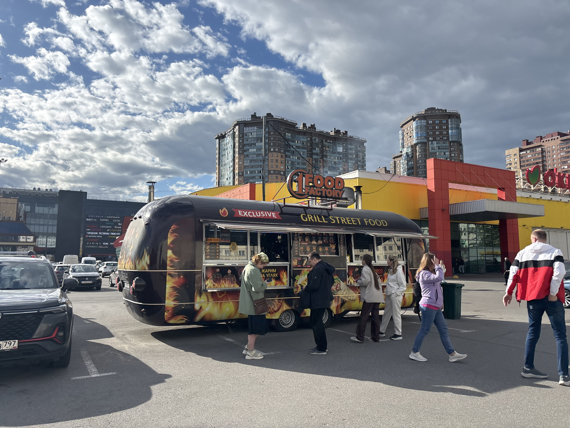 The image shows a "Food Factory" grill street food truck parked in what appears to be a parking lot. Several people are gathered around the truck, some seemingly ordering or waiting for food. A woman in a light purple hoodie walks away from the truck, while a man in a red and white jacket walks towards the right side of the frame. Behind the food truck is a large yellow and red building, likely a shopping center or department store. In the background, there are several high-rise apartment buildings. There are several cars parked in the parking lot and overcast sky. The OCR text indicates the food truck is "exclusive" and sells food grilled over charcoal. The OCR text also identifies several businesses including “Kinopolis” which is a movie theater, suggesting the location is Saint Petersburg, Russia.