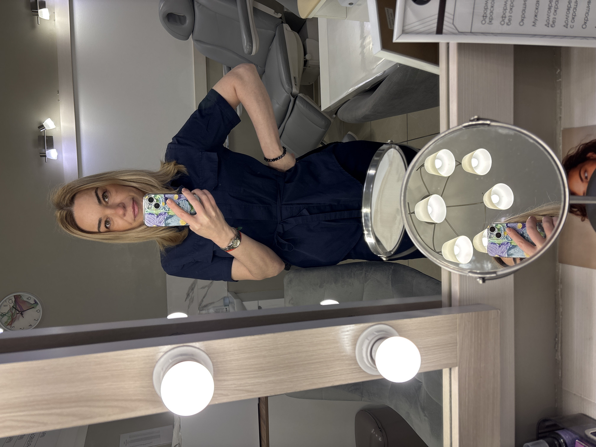The image shows a blonde woman in a dark blue shirt and black pants taking a mirror selfie in what appears to be a beauty salon or cosmetic treatment room. She is standing in front of a large mirror with lights around the edge. A smaller, lighted vanity mirror is visible to her right. A dental-style chair can be seen in the background. The environment suggests a space for cosmetic procedures or beauty treatments, with professional equipment and a neat appearance.
