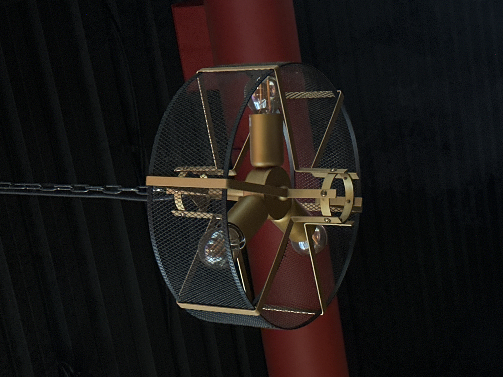 The image features a decorative light fixture suspended from a chain. The fixture consists of a circular wire mesh enclosure with a golden framework. Inside the enclosure, three exposed light bulbs are mounted on a central golden-colored hub. Behind the fixture, a red cylindrical structure is visible. The background is dark and textured. The image was taken in Votkinsk, Russia.