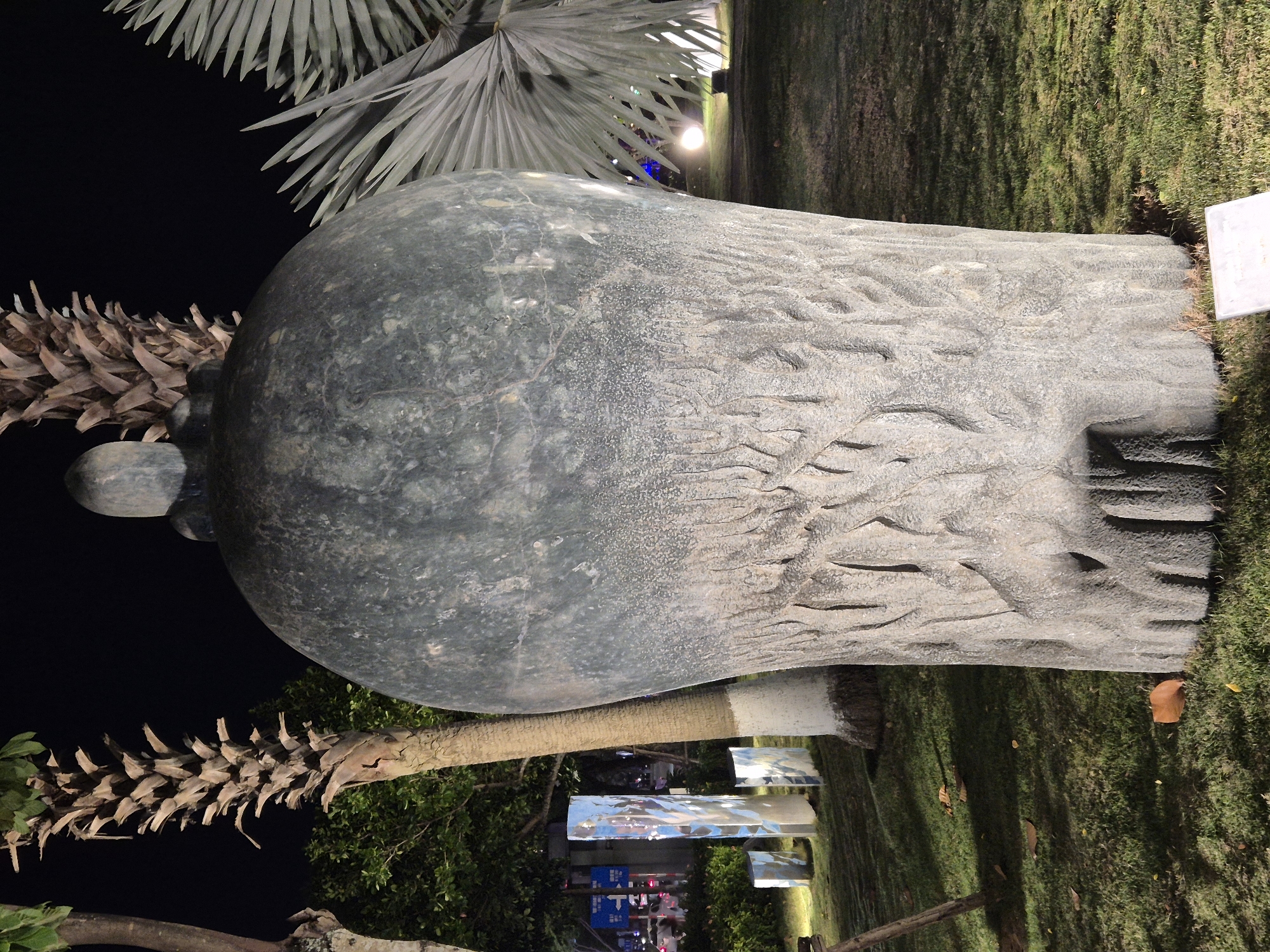 The image captures a nighttime scene in the city of Đà Nẵng, Vietnam, featuring a large, textured stone sculpture as the central subject. The sculpture has a bulbous, vase-like form, with the lower half exhibiting a rough, carved texture resembling tree bark or roots, while the upper portion is smoother and darker. Atop the sculpture is a small, rounded element, giving it a somewhat anthropomorphic or organic appearance.
Surrounding the sculpture is a well-maintained grassy area, illuminated by artificial lighting that casts shadows and highlights the textures of the sculpture and the surrounding foliage. To the left of the main sculpture, several abstract, vertical reflective art installations are visible, glowing with internal light and reflecting the surrounding environment. Palm trees with dry, frond-like leaves are strategically placed around the scene, adding to the tropical ambiance. In the background, glimpses of city lights and traffic can be seen, indicating the sculpture is located in an urban park or public space. The overall atmosphere is serene and artistic, designed to be appreciated after dark.