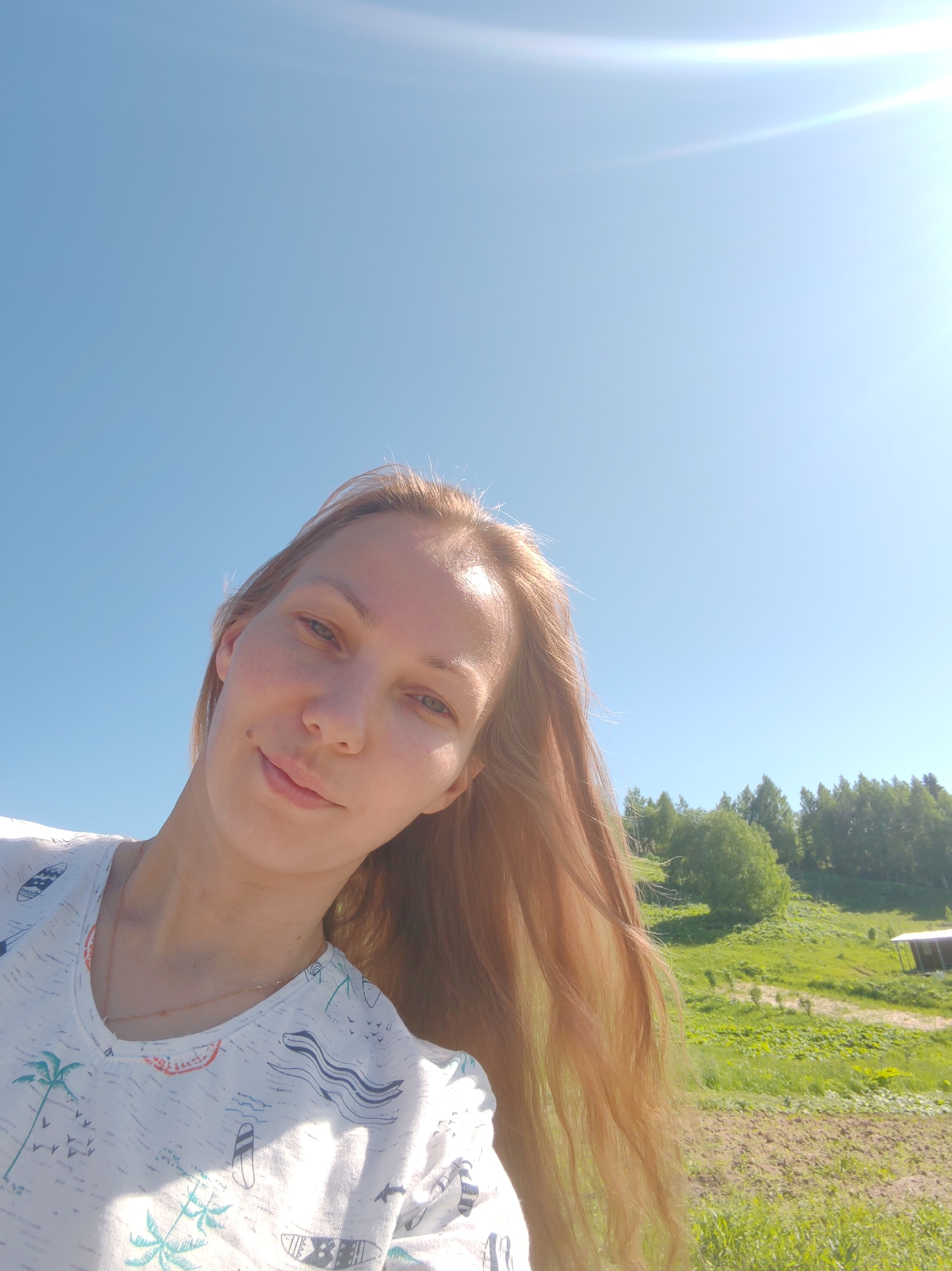 An outdoor, upward-angle selfie-style photograph depicts a young adult female under a clear, bright blue sky in the rural landscape of Чухлэм, Russia. The female, positioned in the lower-left to central frame, has fair skin, light-colored eyes, and long, light brown hair flowing over her right shoulder. She wears a white V-neck t-shirt adorned with tropical patterns including palm trees, surfboards, and waves, and a delicate gold-colored chain necklace. Her head is slightly tilted, and she looks directly at the camera with a subtle, closed-mouth smile. A small mole is visible on her upper lip. Strong sunlight from the upper right casts a lens flare and illuminates the right side of her face and hair. The background features a verdant rural environment with rolling green hills, dense trees along a distant ridge, and a visible dirt path. A small, light-colored building with a dark roof is partially visible in the mid-ground on the far right. The foreground on the right shows sparsely vegetated ground.