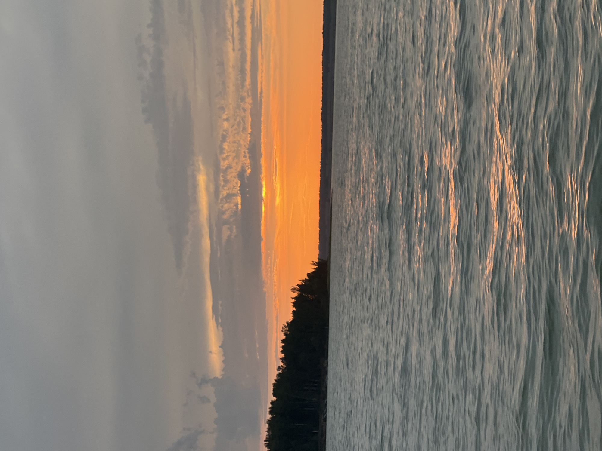 The image shows a body of water with a tree line on the horizon. The sky is filled with clouds and hues of orange and grey, indicating a sunset or sunrise. The water is calm with gentle ripples reflecting the sky's colors. The location is identified as Beregovoe, Russia. There are no people or animals visible in the image.