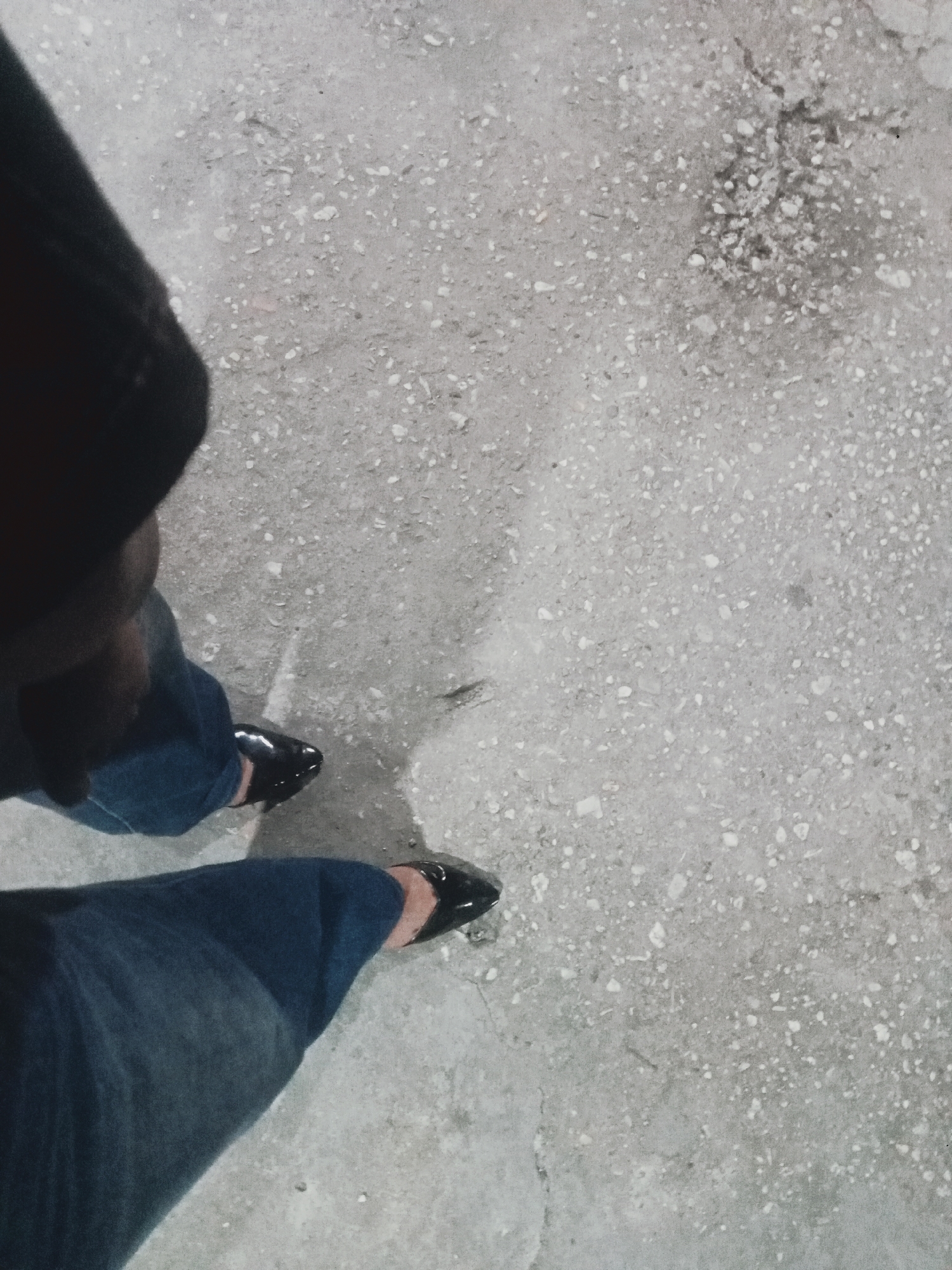 The image shows a first-person perspective of someone standing on a speckled, gray concrete surface. The subject is wearing dark blue jeans that are rolled up at the cuffs and shiny black pointed-toe shoes. To the left of the frame, a dark shadow is cast, obscuring part of the subject's upper body and a portion of the concrete. The concrete itself appears to be somewhat uneven and textured, with visible small stones or aggregates embedded within it. There is no clear indication of the specific location within Bumbung, Indonesia, beyond the material of the ground. The lighting suggests it could be daytime, possibly overcast, or artificial lighting at night.