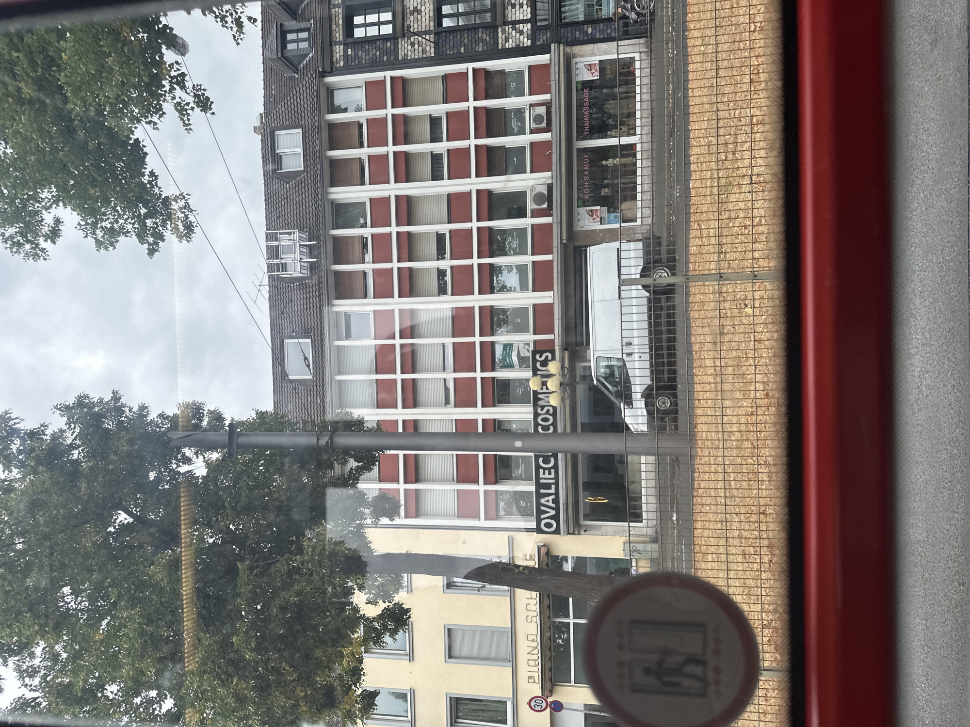 The image captures a street scene in Krefeld, Germany, viewed from inside a vehicle. In the foreground, a red band and textured surface suggest the interior of a bus or tram. A circular sign with a pictogram of a person exiting a doorway is visible on the left. Beyond the glass, a row of buildings lines the street. The building on the far left is pale yellow, featuring windows and a sign that reads "PIANO SCHULE" (Piano School). Adjacent to it is a building with a striking red and white facade, featuring numerous grid-like window frames. A large sign on this building says "OVALIEC COSMETICS". Further to the right, a white van is parked in front of a shop displaying signs for "KOH SAMUI THAIMASSAGE". The street is bordered by a metal fence and a strip of ground covered with dry leaves or wood chips. Trees with green foliage are present on the left and right edges of the frame, with a utility pole situated between the buildings. The sky above is overcast.