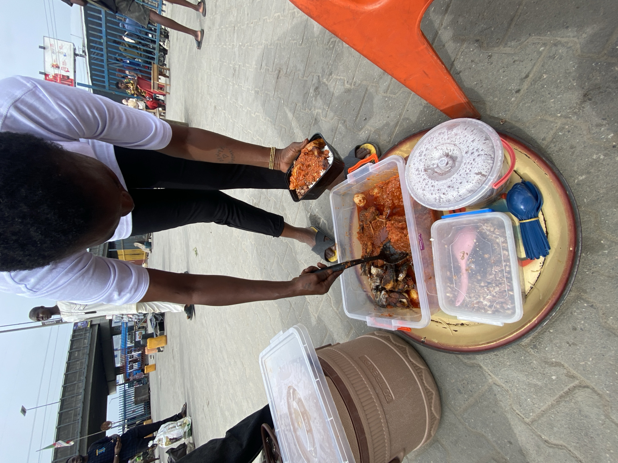 An overhead medium shot captures a street vendor in Lagos, Nigeria serving food. The vendor, dressed in a white shirt and black pants, is holding a black takeout container filled with rice and stew in their left hand. With their right hand, they are using a black spoon to scoop fish from a clear plastic container filled with stew onto the rice. This container, along with a white bucket of rice and another clear plastic container, is resting on a round gold tray. An orange plastic chair is partially visible in the upper right corner.
The ground is paved. In the background, there is a busy street scene with pedestrians, vehicles, and an overpass. People are standing and walking around in the distance, and there's a glimpse of an overpass structure in the upper left. Some individuals appear to be involved in various activities, contributing to the hustle and bustle of the Lagos street. A Nigerian flag is displayed in the background.