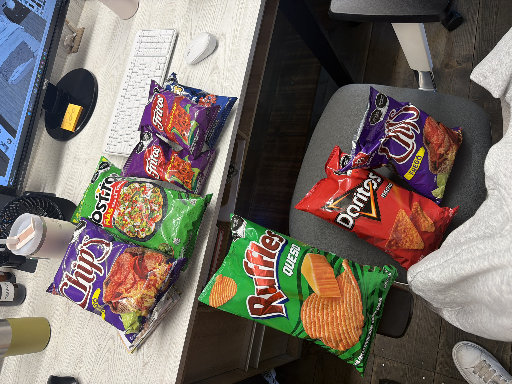 The image shows a desk and chair covered with various bags of chips. On the desk are bags of Sabritas Chips (Chile y Limón Ardiente flavor), Fritos Flamin' Hot, and Sabritas Ostito (Salsa Verde flavor). Also on the desk are a computer monitor, keyboard, mouse, pens, and a container. On the chair are a bag of Doritos Nacho chips, Barcel Fuego chips, and a bag of Ruffles Queso chips. The photo was most likely taken indoors, possibly in a home or office setting. The items appear to be arranged somewhat haphazardly. Puebla, Mexico is not mentioned in the image.