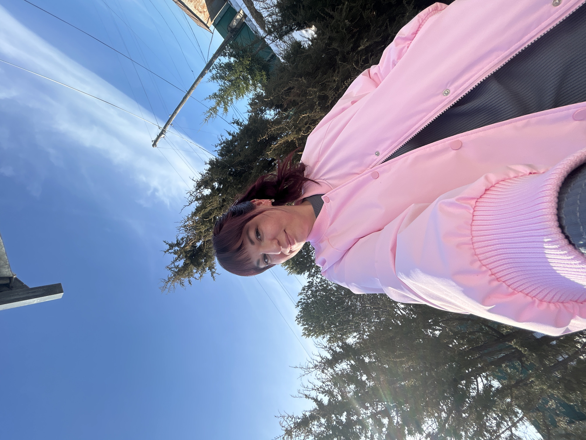 A woman wearing a pink jacket is outside in Фоки, Russia. Trees are visible in the background, and a clear blue sky with some clouds is overhead. Power lines are strung across the sky.