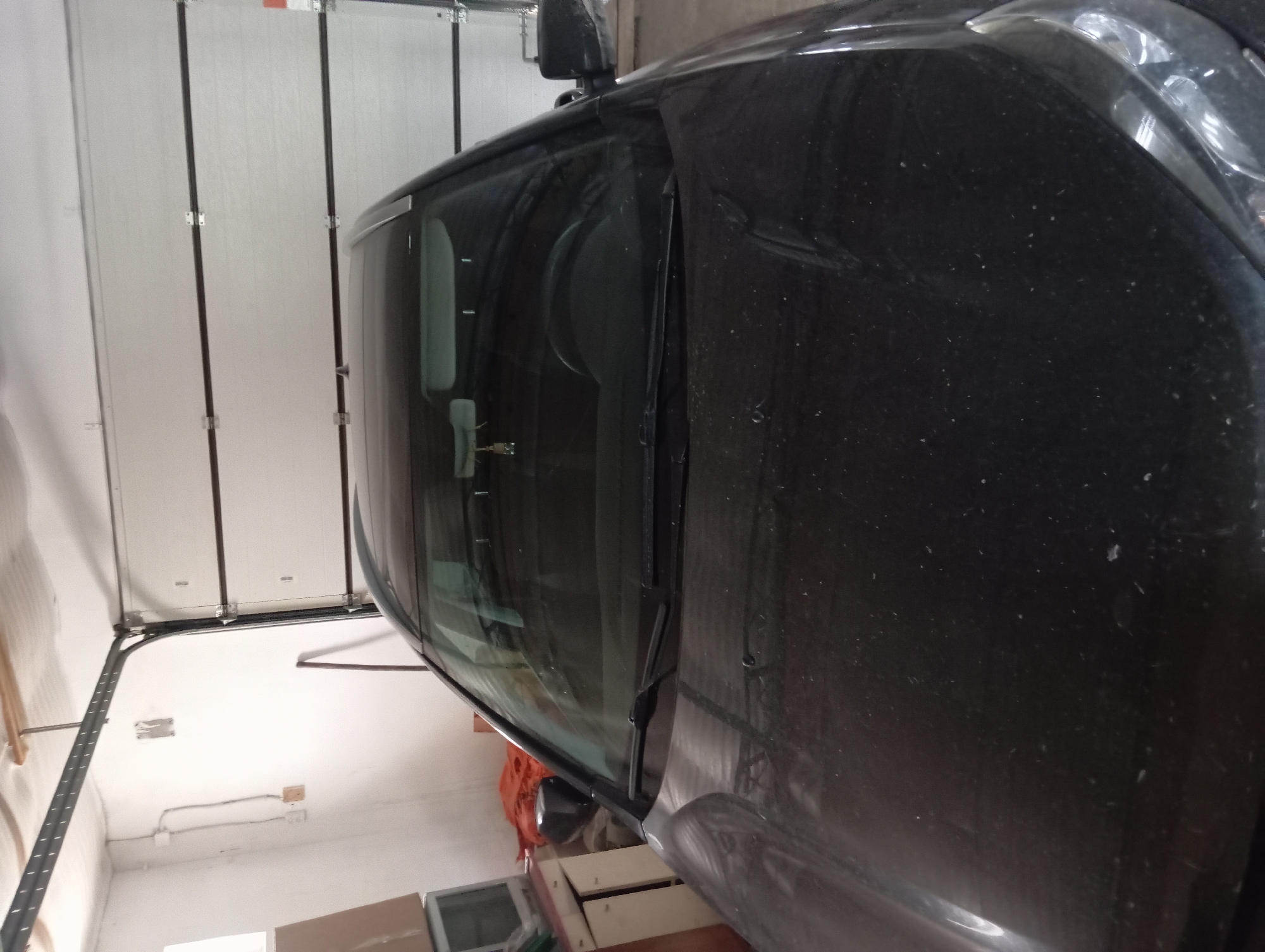 A black car is parked inside a garage in Peredovaya, Russia. The car is facing forward, with its hood and windshield prominently visible. The garage door is open, revealing a white sectional door in the background. The garage appears to be dimly lit, with some clutter visible to the left of the car, including shelves and what looks like an old television. The car's hood is covered in dust.