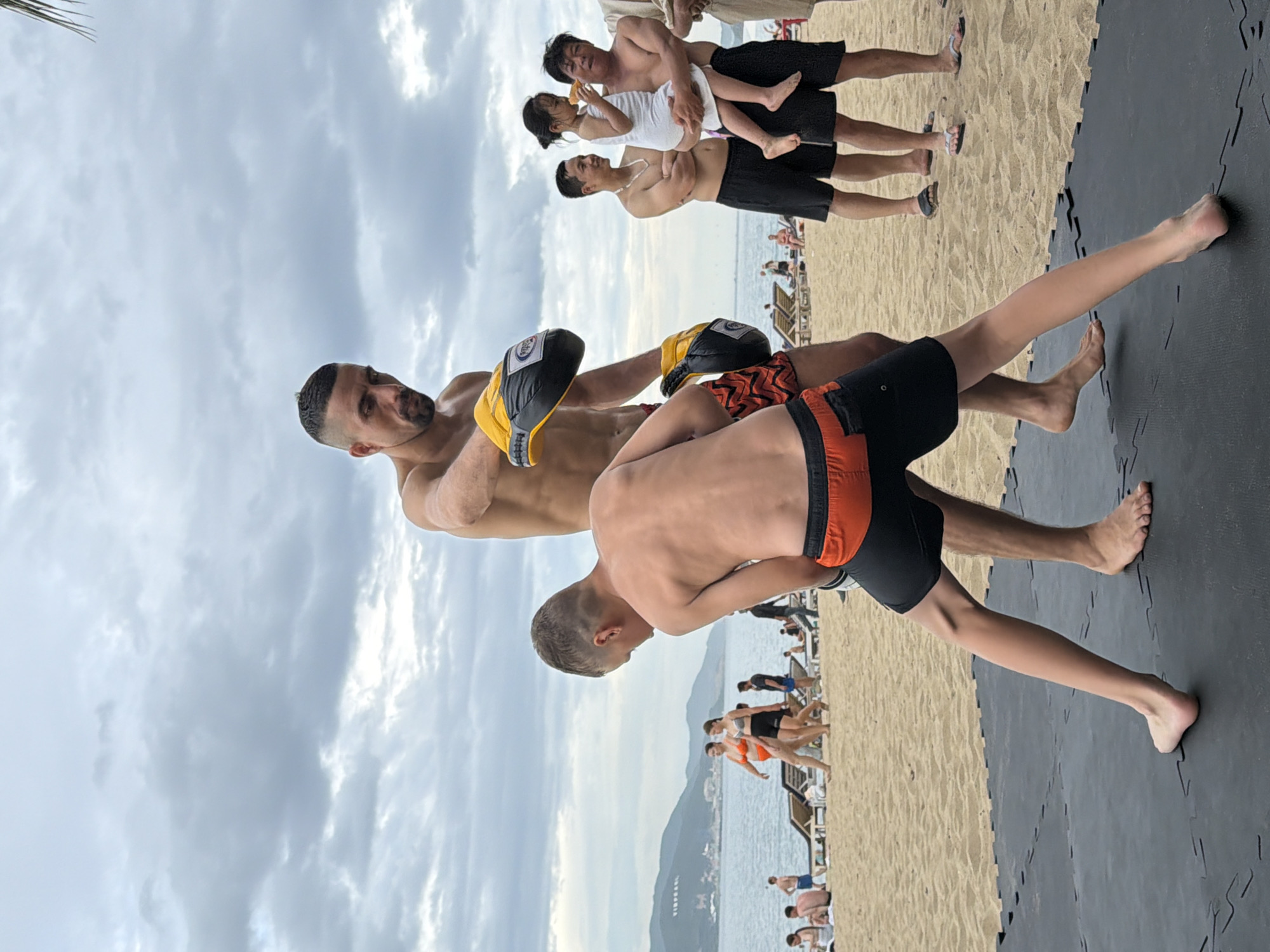 This image depicts an outdoor training session on a beach, likely in Nha Trang, Vietnam, given the presence of palm trees and the distinctive mountainous coastline. In the foreground, two individuals, a younger male and an adult male, are engaged in what appears to be martial arts training. The younger male, wearing black shorts with an orange band, is in a defensive stance on a black mat. The adult male, shirtless and wearing black shorts, is wearing boxing gloves, one of which is extended. He has a beard and a buzz cut.
In the background, the beach is populated with other people. Several individuals are seated on beach chairs, and others are walking along the shoreline. The sea is visible, with a coastline featuring green hills and some buildings. The sky is overcast with scattered clouds. Further back on the beach, a group of people includes a man holding a young child who is eating something. Other men are also present in this background group. The overall scene suggests a casual, recreational environment where physical activity is taking place alongside general beach-going activities.