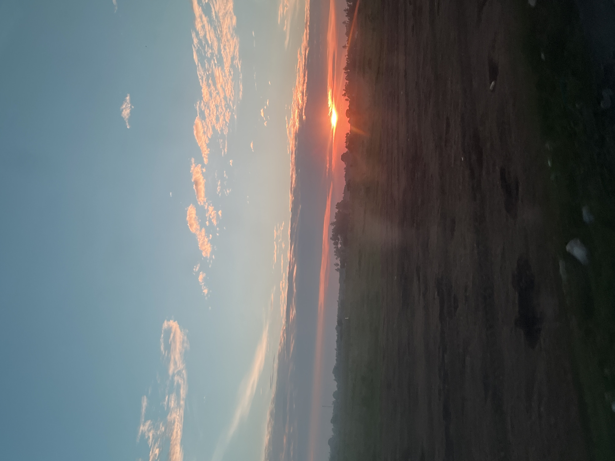 A low-angle, vertical photograph captures a sunset over a rural landscape in Meunasah Pupu, Indonesia. The sky transitions from a clear, pale blue at the top to hues of orange and red near the horizon, illuminated by the setting sun. Wispy clouds, tinged with pink and orange, are scattered across the sky.
The foreground consists of dark, uneven terrain with patches of dry earth and sparse vegetation, suggesting a field or undeveloped land. The middle ground is a hazy expanse of green, punctuated by silhouetted trees and structures, indicative of a distant village or agricultural area. The sun, a bright orb, is partially obscured by clouds and trees on the horizon, casting a warm glow and light rays across the scene. The overall atmosphere is serene and natural.