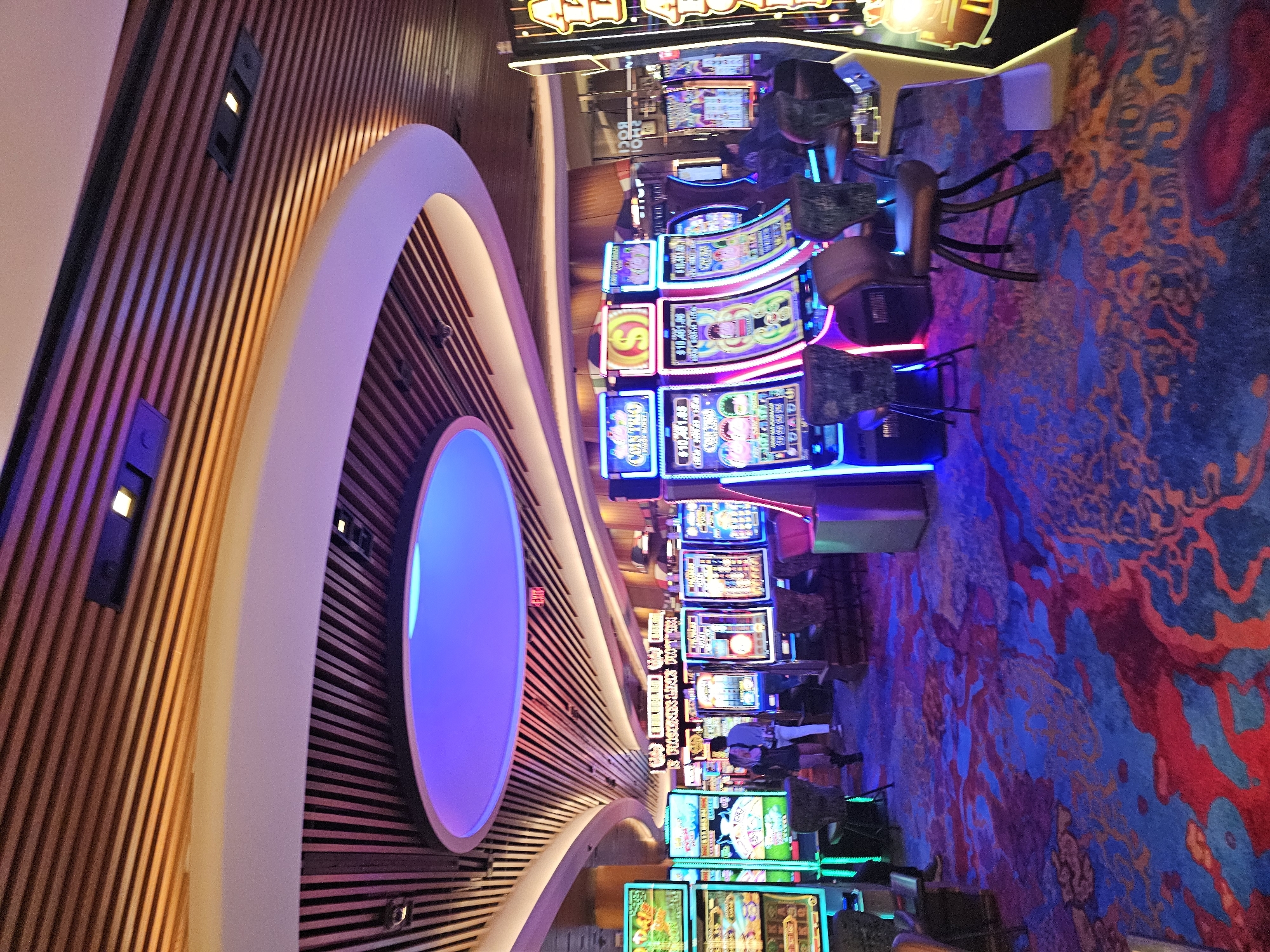 The image shows an interior view of a casino on the Hollywood Seminole Reservation in the United States of America. The scene is dominated by rows of slot machines with illuminated displays and neon accents. A patterned carpet in shades of blue, orange, and red covers the floor. Along the left side is a curved architectural feature with wooden slats and a circular illuminated element. Several people are visible, either standing or sitting at the slot machines. The overall lighting is vibrant and colorful, creating a lively atmosphere.