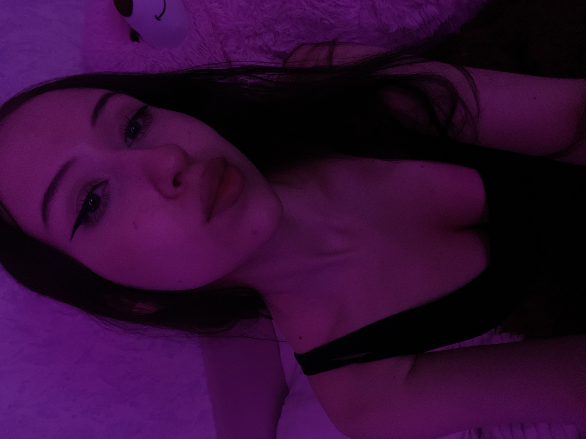 The image is a close-up selfie of a young woman with long dark hair. She is wearing a black tank top. The lighting is a vibrant purple hue. To her right, a portion of a large white teddy bear is visible. The background appears to be a textured surface, possibly a blanket or wall, also illuminated by the purple light. No specific location in Votkinsk, Russia can be determined from the image.