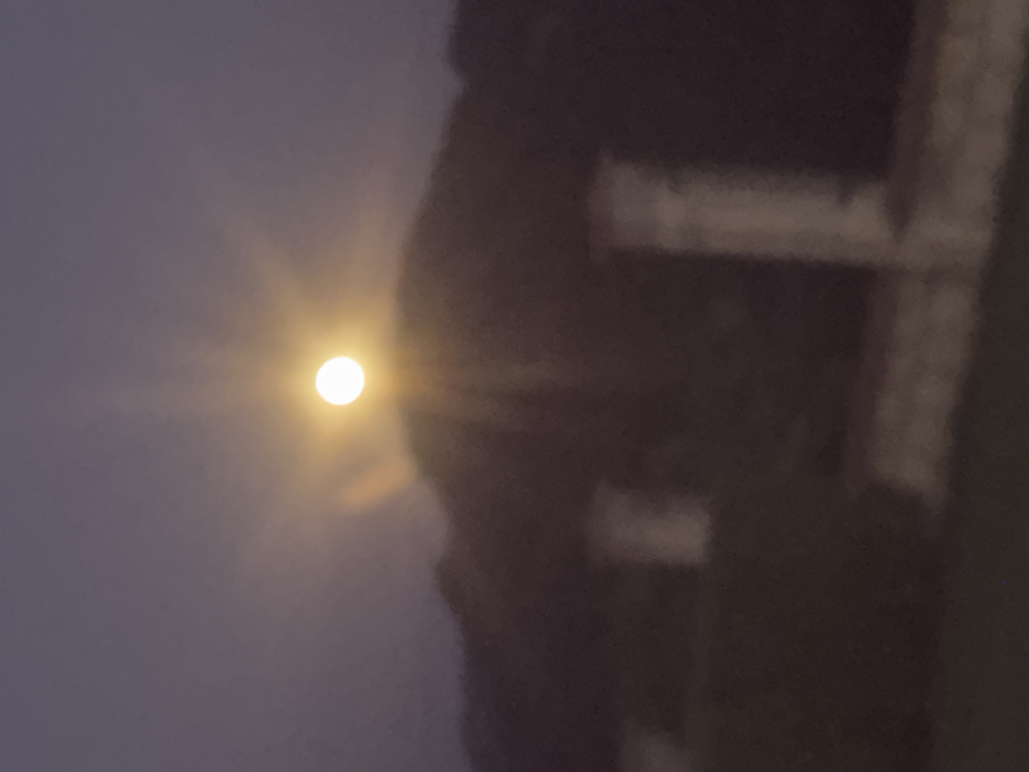 The image depicts a full moon rising behind a silhouetted mountain range. The moon is a bright, luminous orb with visible rays emanating from it, suggesting a clear or slightly hazy sky. Below the mountain range, indistinct structures, possibly buildings or remnants of buildings, are visible in the dim light. The foreground appears to be a dark, shadowed area, possibly land or vegetation. The overall scene evokes a sense of tranquility and natural beauty. The location is not specified in the image.