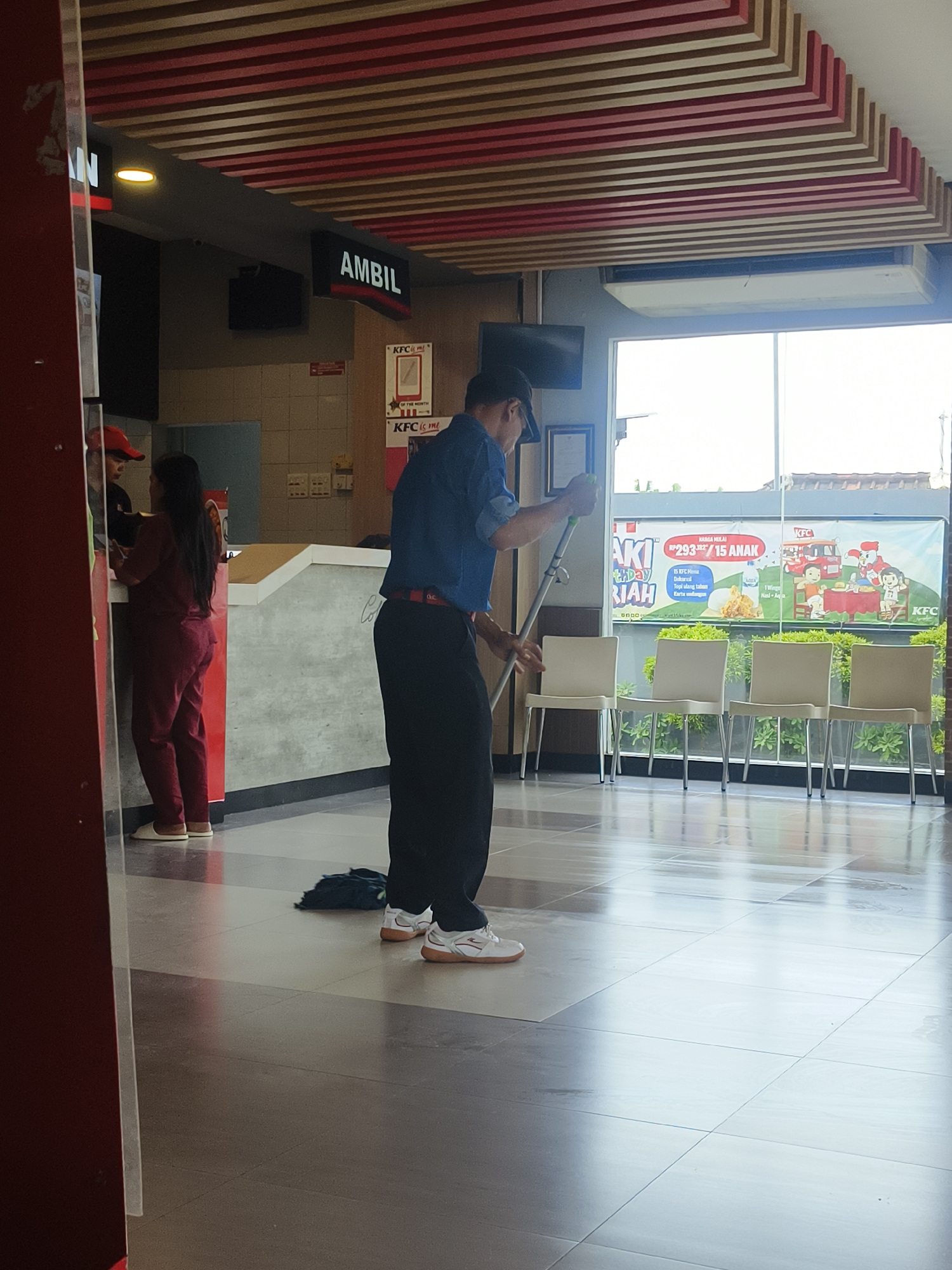 The image shows the interior of a KFC restaurant, likely located in Denpasar, Indonesia, given the Indonesian language on the signage. In the foreground, a man in a blue shirt and black pants is mopping the floor with a blue mop. He is wearing white sneakers. To his left, two employees are visible behind a counter. One is wearing a red uniform with matching pants and is interacting with another person whose face is not fully visible. The background features a large window overlooking an outdoor area with trees. Various KFC promotional banners are displayed behind the counter and on the window. The ceiling has a distinctive slatted design with red and wood tones. The overall environment appears to be clean and functional, consistent with a fast-food establishment.