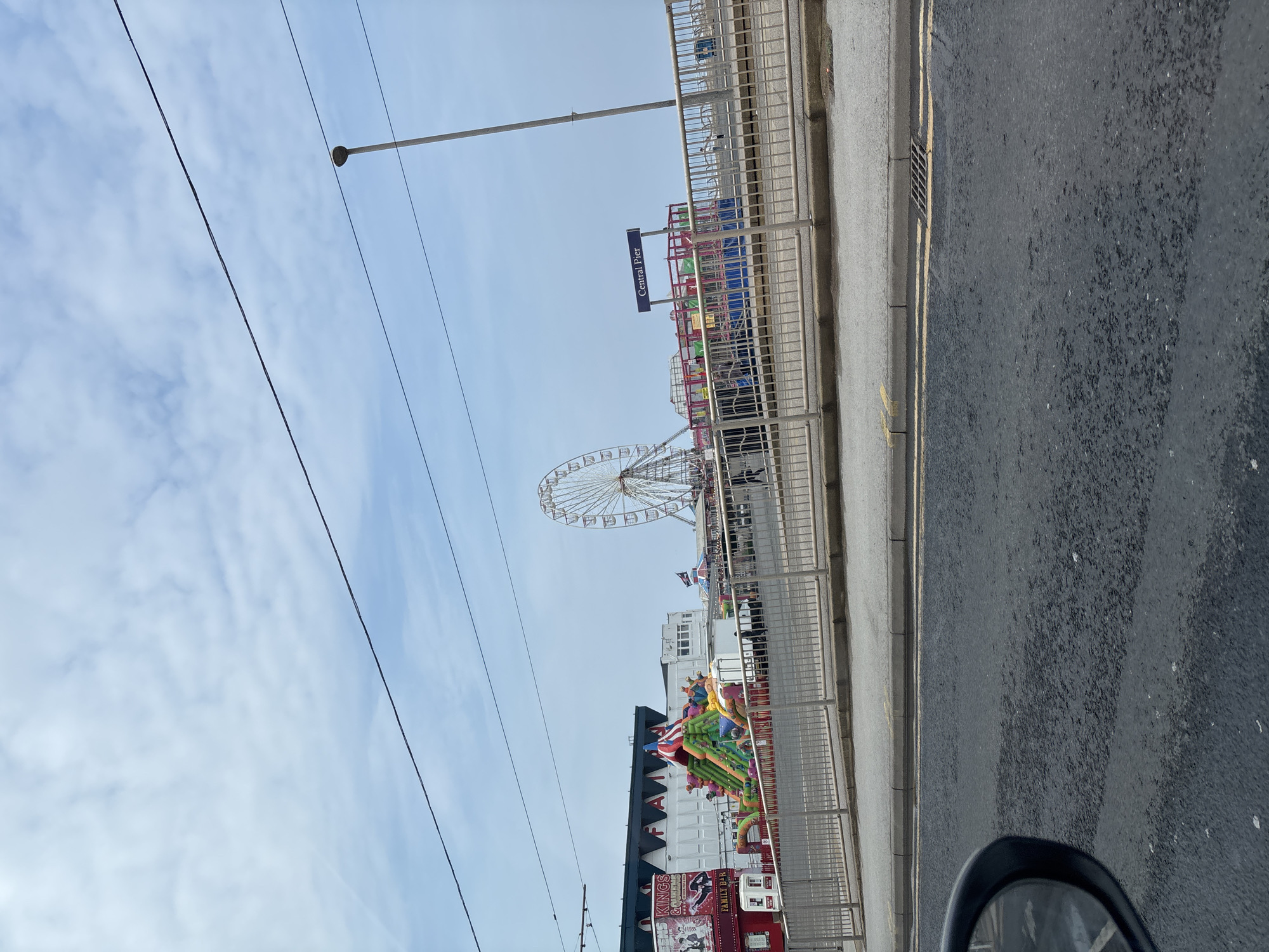 The image shows a street scene in Blackpool, United Kingdom, with the Central Pier visible in the background. A large Ferris wheel stands prominently near the pier. Other amusement park attractions are visible, including brightly colored rides and stalls. A street runs along the right edge of the image. The sky is pale blue with light clouds. Power lines are strung overhead. A lamppost is visible. A roadside barrier separates the sidewalk from the amusement park attractions. A street sign indicating “Central Pier” is visible. The image appears to be taken from a vehicle, with a portion of the vehicle's side mirror visible in the lower right corner.