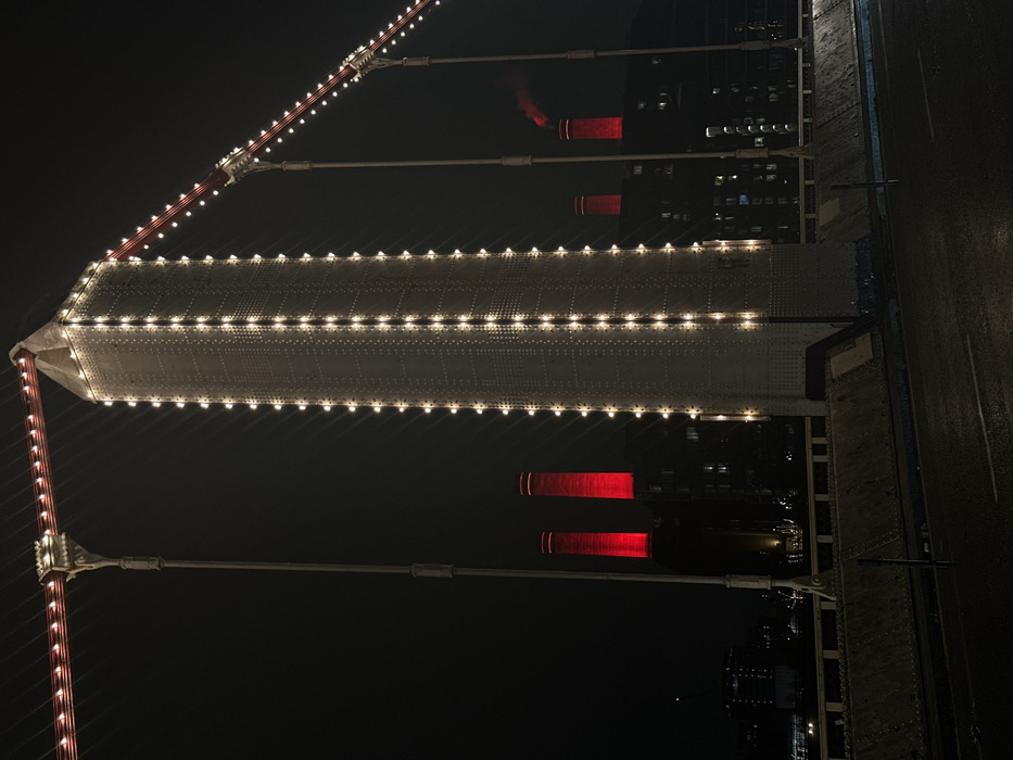 An elevated, nighttime view of a bridge structure. The bridge's tower, adorned with rows of lights, extends horizontally. Red lights are visible on distant buildings beneath the bridge's cables. The bridge deck can be seen along the right edge of the image, with a wet, reflective surface. The dominant color is black, highlighting the bridge's lit elements against the dark sky.