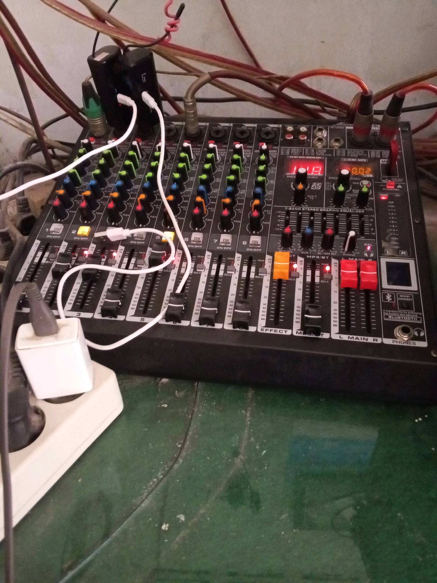 This is a close-up, high-angle shot of a black audio mixer with numerous sliders, knobs, and buttons, set against a backdrop of tangled cables and a dusty green surface. The mixer is the central subject, featuring channels labeled with numbers, "GAIN," "EQ," "AUX," "PAN," and "ON/OFF." Some sliders are set at various levels, indicated by the white markers. Several red and orange slider caps are also visible. A small digital display shows "002." A white USB cable connects to a black power adapter plugged into a white power strip, which is situated on the left side of the frame. The cables are a mix of red, brown, and black, and appear somewhat disorganized. The environment appears to be an indoor setting, likely a home or a small venue, given the presence of domestic electrical outlets and the general condition of the surfaces. The specific location in Deli Tua Timur, Indonesia, is not explicitly depicted in the image itself but is provided as context for the scene.