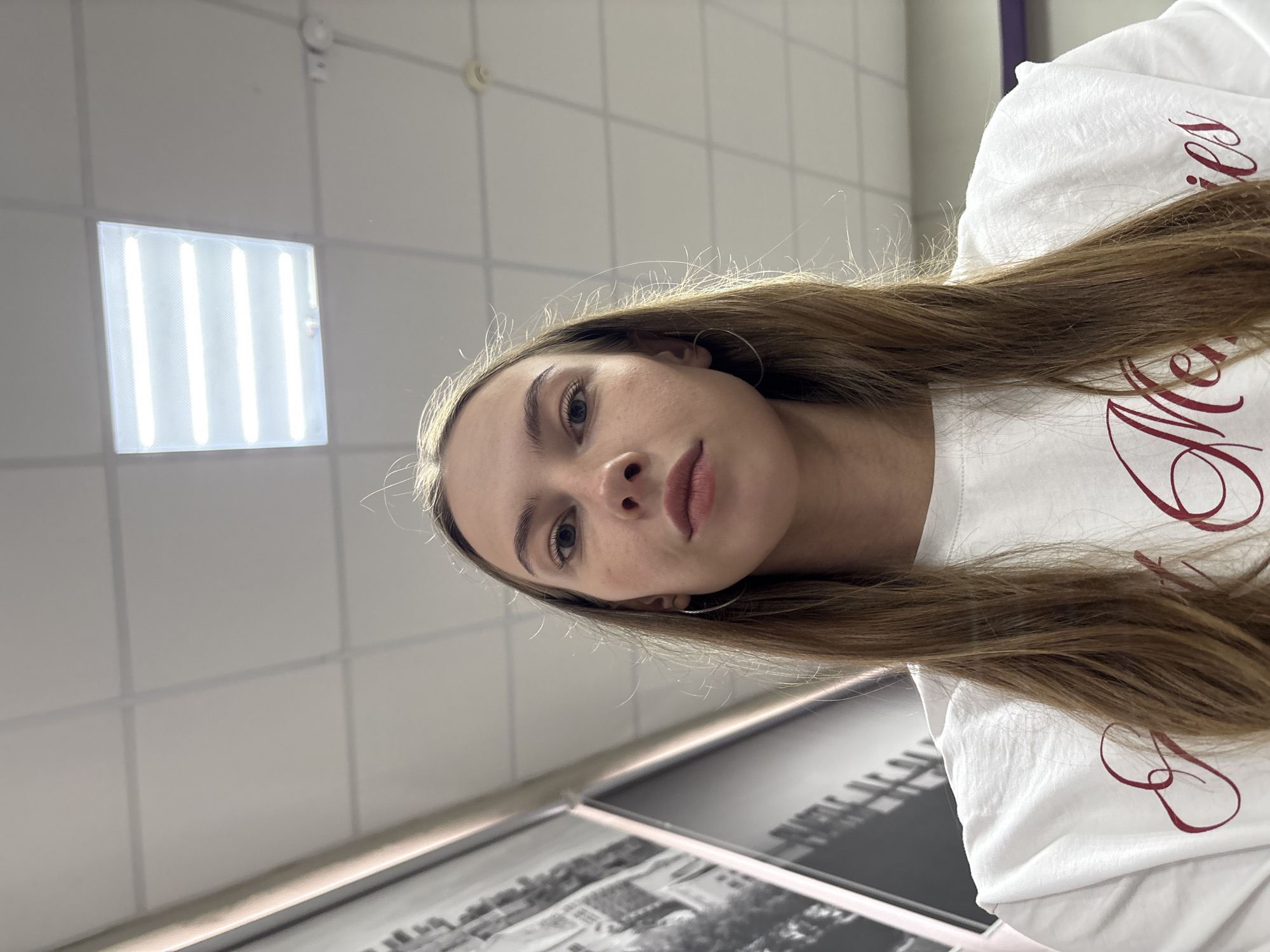 The image is a low-angle, chest-up selfie of a young woman with long, straight, brown hair. She is wearing a white t-shirt with red script lettering that reads "Sweet Memories". Her eyes are open and she is looking directly at the camera. She has large hoop earrings in both ears. The background consists of a white ceiling with a grid pattern and a rectangular fluorescent light fixture. To the left of the woman is a large black and white photograph or mural depicting buildings and possibly trees. The lighting suggests an indoor environment. There is no specific visual information to indicate the location is in the city of Perm, Russia.