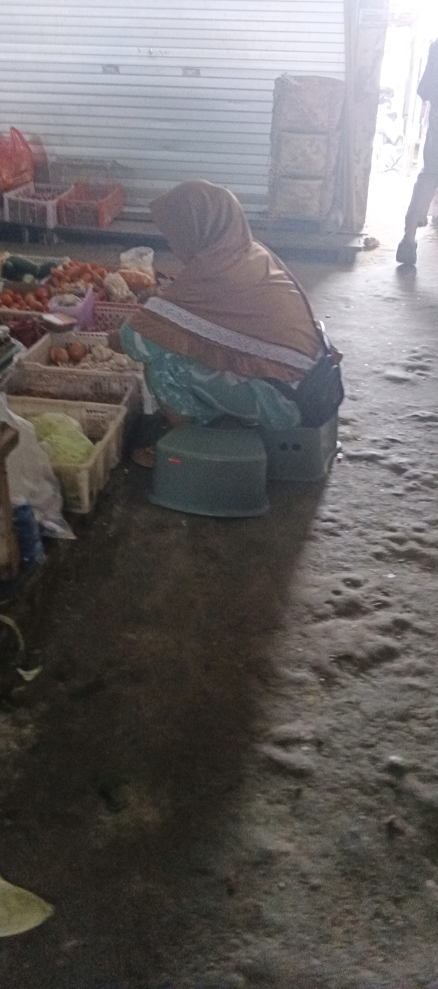 The image depicts a market scene in Sekaran, Indonesia. A person wearing a light brown hijab is seated on two stacked green plastic stools in the foreground, facing away from the camera. The person's right arm is bent, and their left arm is obscured. They are covered by a light blue and green fabric. In front of the seated person, a variety of produce is displayed in crates. These include leafy green vegetables, tomatoes, and other unidentifiable fruits and vegetables. To the left of the crates, a red plastic bag is visible. In the background, a large roll-up door, partially open, reveals a bright, possibly outdoor area. To the right, a person is standing, wearing dark pants and a light-colored shirt, with their back to the camera. The ground is wet and appears to be concrete or asphalt, with visible puddles and a rough texture. The overall lighting suggests it is daytime, with diffused light entering from the open doorway.