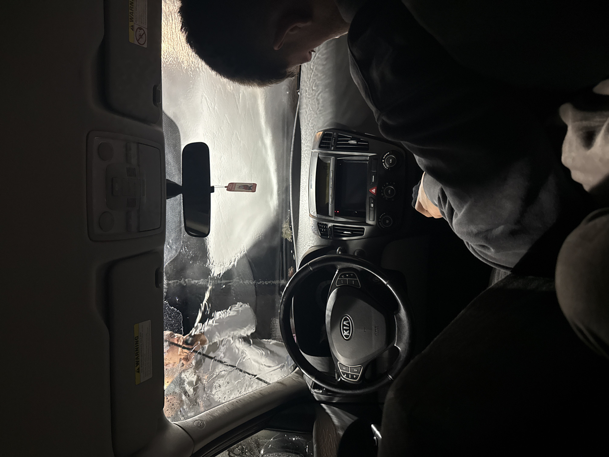 The image shows the interior of a car, likely a Kia, during a car wash. Water streams down the front windshield. Two people are visible: one outside the car cleaning the windshield with a brush and the other, presumably the driver, is inside. The dashboard and steering wheel are in focus. The location in Blagoevgrad, Bulgaria cannot be determined from the image.