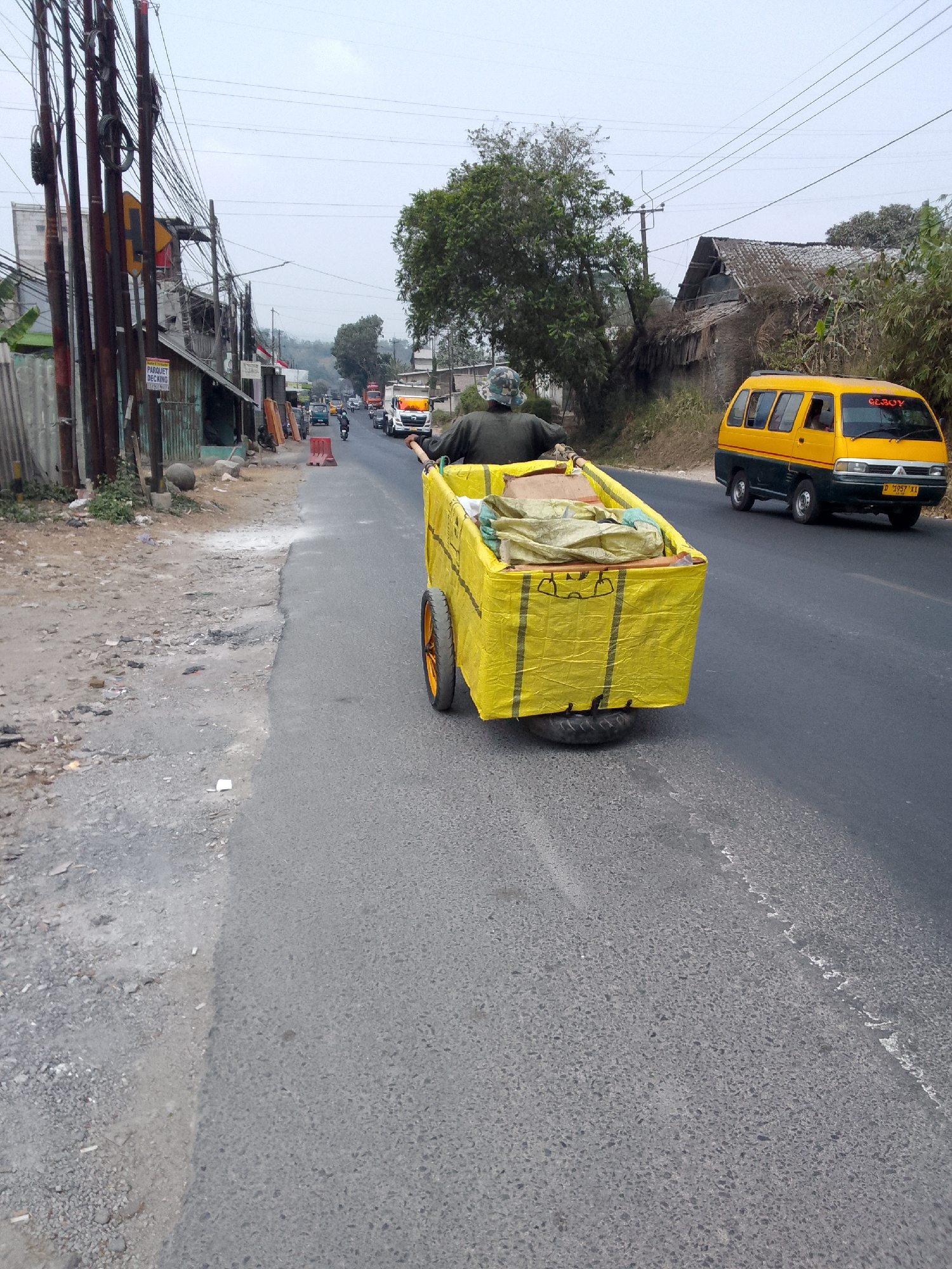 A person wearing a camouflage hat and dark clothing is pushing a large, yellow, rectangular cart with two orange wheels on a paved road. The cart is covered with a yellow tarp, and appears to contain some bundled items. The person is positioned at the back of the cart, holding onto a handle. The scene is set in an outdoor environment with a road running through it. On the left side of the road, there are several utility poles with numerous wires, some buildings with corrugated metal roofs, and a dusty, uneven ground area with debris. On the right side of the road, there is a yellow and dark green van with a license plate that reads "D 7957 XI". In the background, there are trees, more buildings, and a hilly landscape under an overcast sky. The overall impression is of a street scene in a town or village.