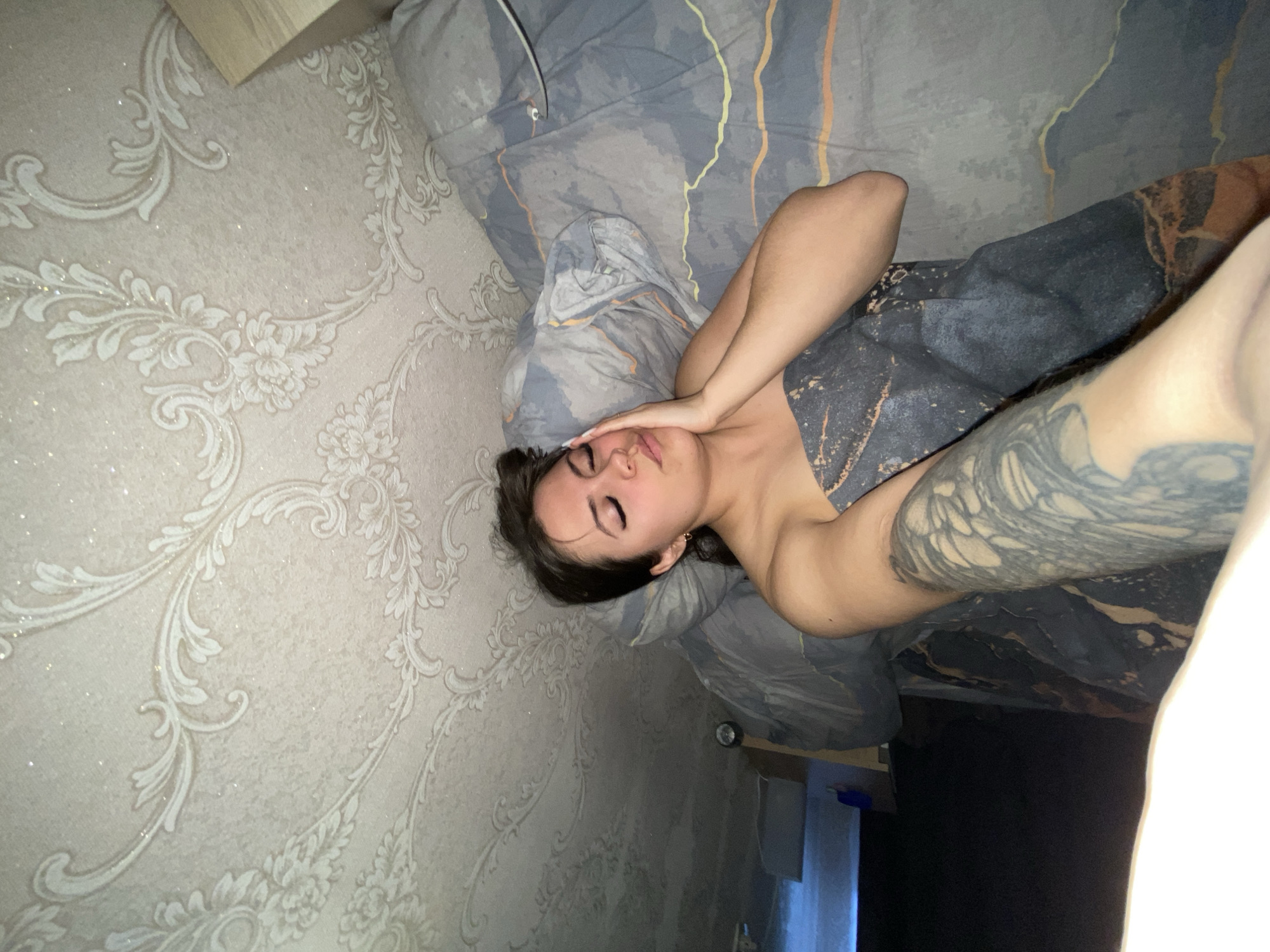 A medium shot, taken from a slightly high angle, depicts a person lying on their side in what appears to be a bed. The image is oriented sideways, with the person's head positioned towards the left and their body extending to the right. The subject has dark hair and is wearing a top with a dark, possibly grey or black, abstract pattern. Their left arm is prominently displayed, covered in intricate black and grey tattoos. The person's right hand is placed over their forehead and eyes, with visible long, light-colored fingernails. Their eyes are closed, and their mouth is slightly open, suggesting a state of relaxation or sleepiness. A small, golden hoop earring is visible on their right ear.
The person is resting on bedding featuring a dark grey or blue base with abstract, fluid lines in shades of orange, yellow, and white. A pillow with a matching design supports their head. The background wall on the left is adorned with light-colored wallpaper featuring an ornate, classical floral and scroll pattern, accented with a subtle glitter effect. A small wooden object, possibly part of furniture, is partially visible in the upper left corner. The indoor setting, likely a bedroom, is situated in Chaikovsky, Russia. There is no visible text in the image.