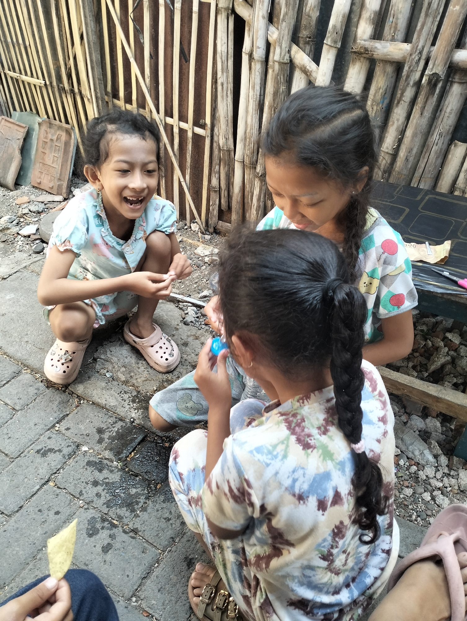 Three young girls are gathered on a paved ground area in what appears to be an outdoor setting. The girl on the left, with dark curly hair, is squatting and laughing. She is wearing a light blue t-shirt with a pattern of cats and what seem to be flowers, and pink clogs. In the center, a girl with dark hair in a braid is leaning forward, looking down at something she is holding. She is wearing a shirt with a colorful geometric pattern and balloons. A third girl, in the foreground and facing away from the camera, also has dark hair in a braid, tied with a pink band. She is wearing a light-colored shirt with a tie-dye pattern of floral motifs. The ground is paved with grey bricks, with some wet patches and debris. In the background, there is a fence made of vertically arranged bamboo stalks. Some broken pottery pieces and rubble are visible near the fence. A dark-colored object, possibly a bench or table, is located to the right of the girls. The overall scene suggests a casual gathering of children in a somewhat unkempt outdoor environment. The location is Situbondo, Indonesia.